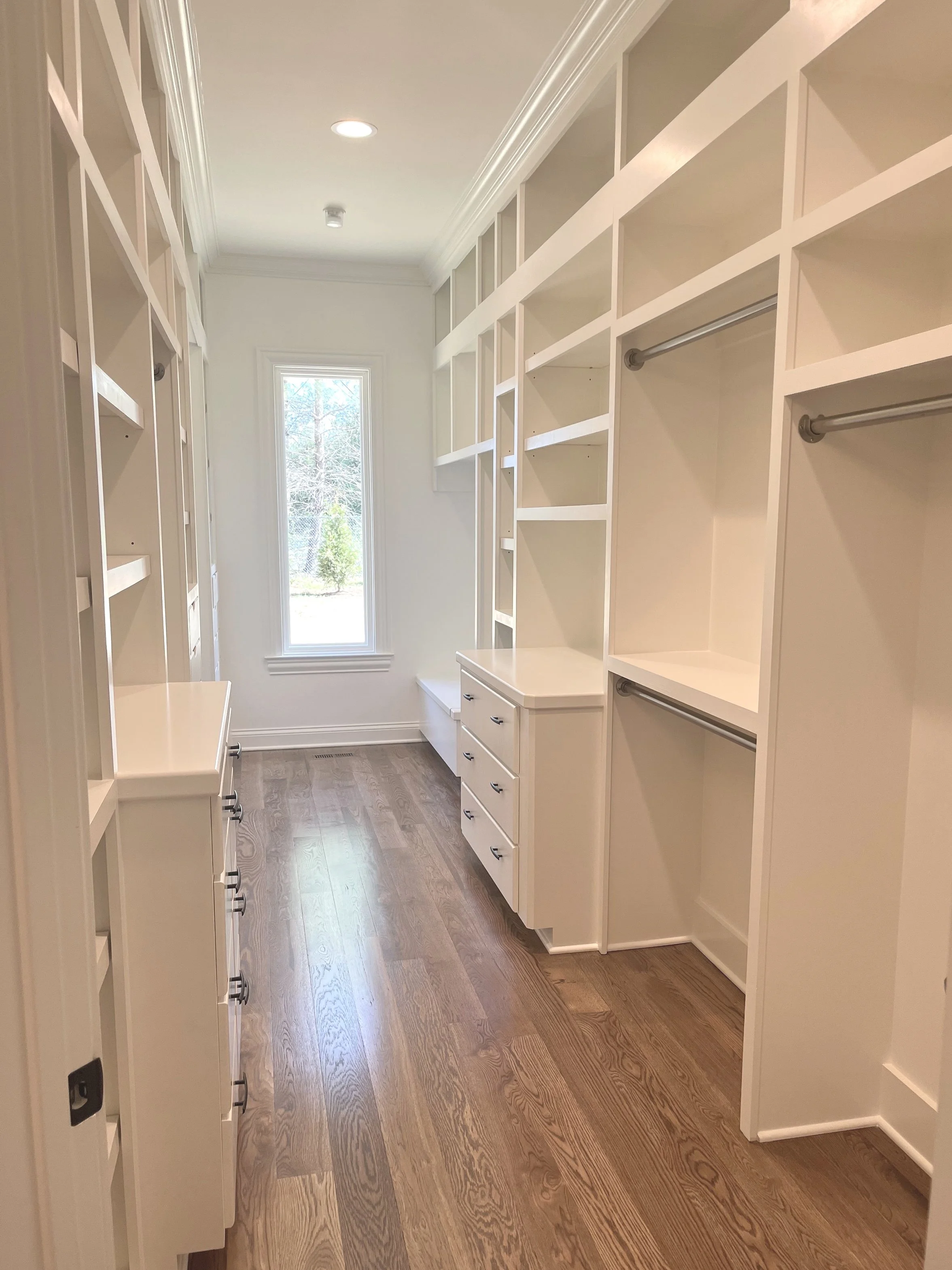 Empty walk-in closet with white built-in shelving, drawers, and hanging rods, wooden flooring, and a window letting in natural light.