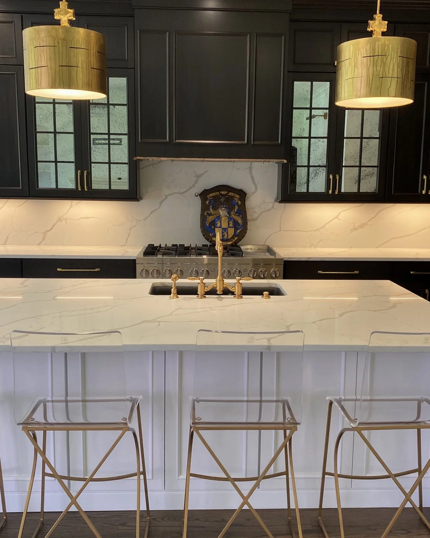 Modern kitchen with black cabinets, white marble countertops, gold fixtures, and clear acrylic bar stools, featuring a double pendant light with wood grain texture and a crest above the stove.