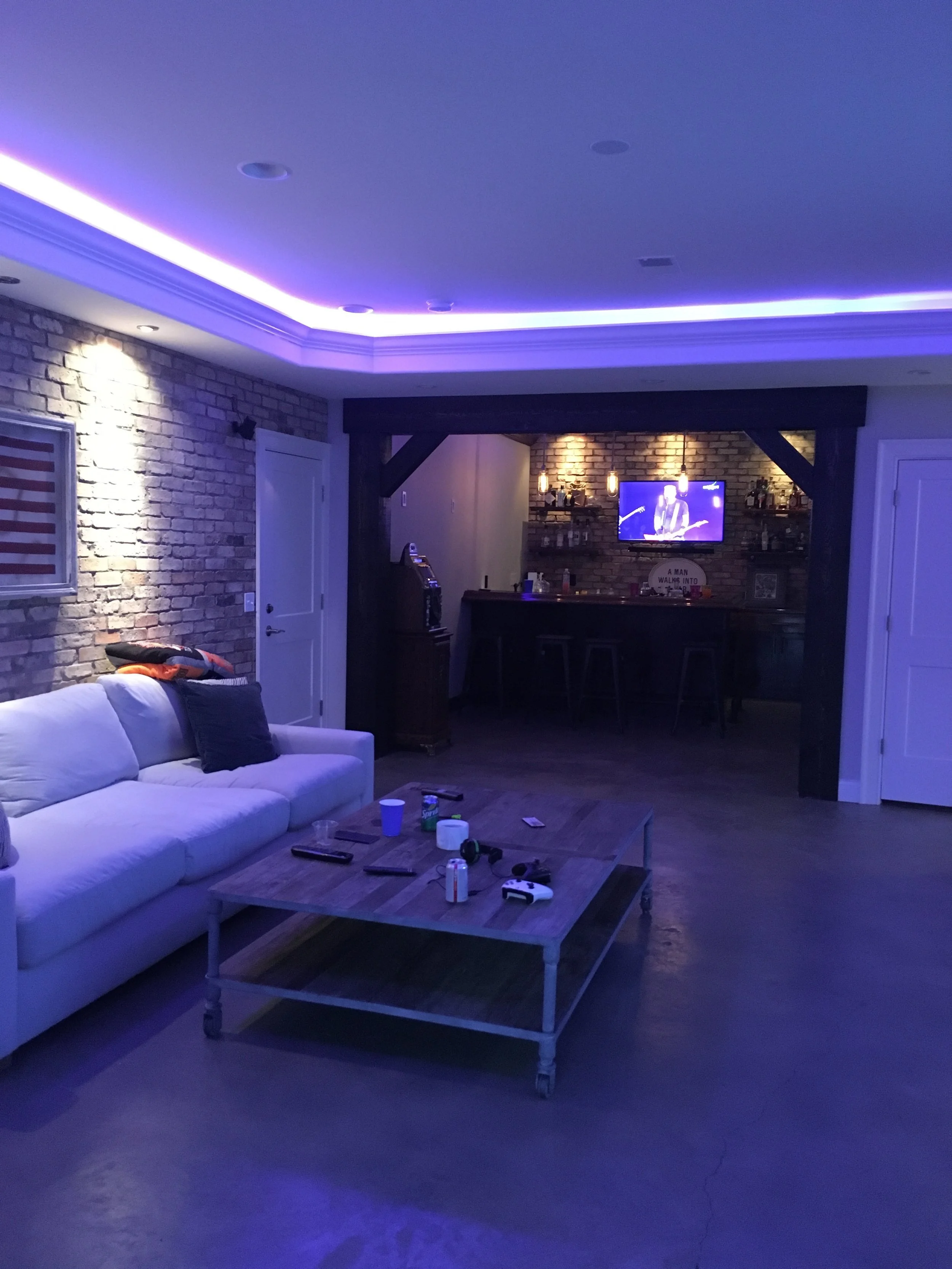 Living room with white sofa, brick walls, wooden coffee table with remote controls and drinks, and a bar area with a television in the background, illuminated with purple LED lighting.
