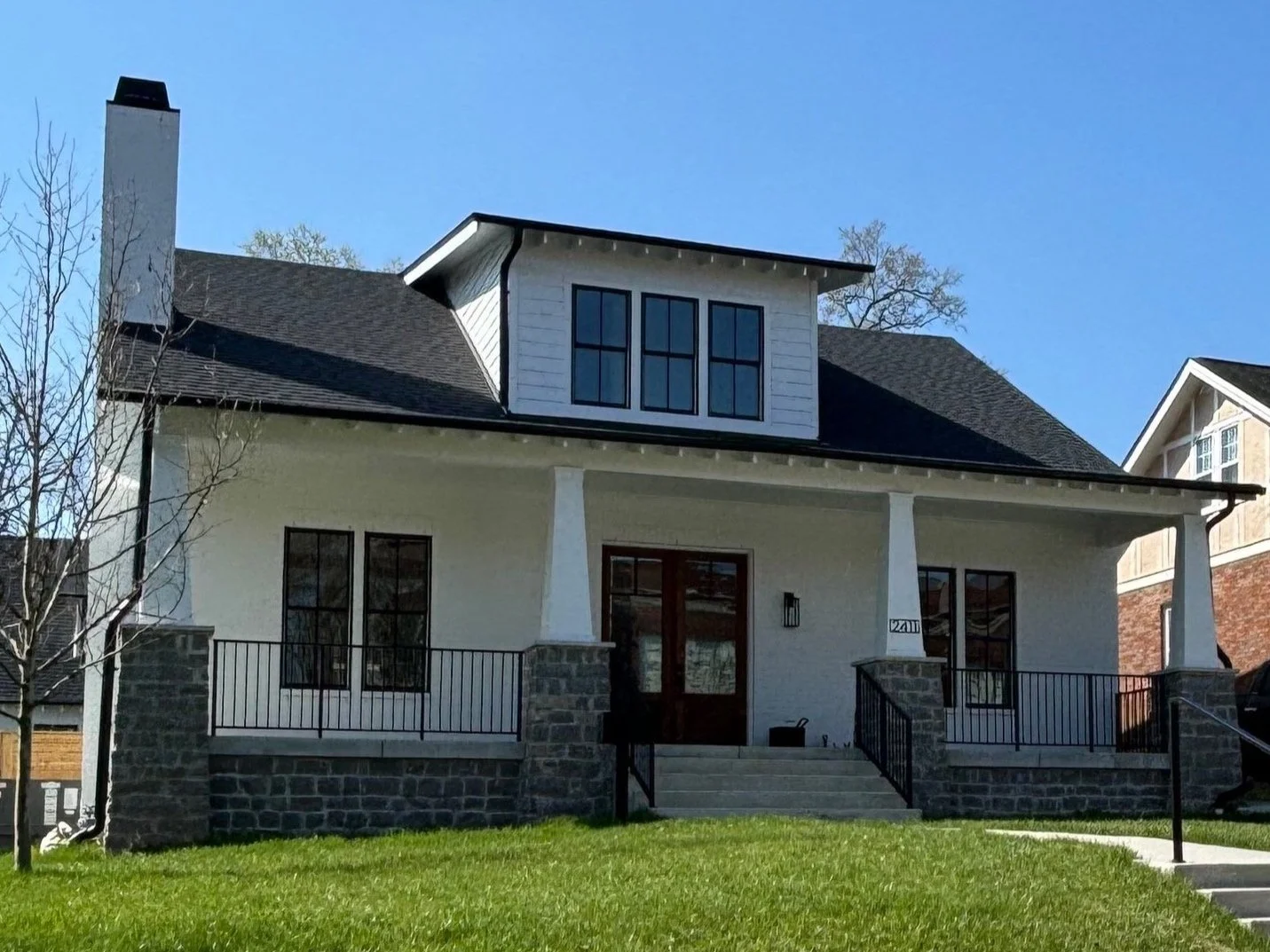 A two-story house with white exterior, black roof, and stone accents, featuring a front porch with steps, black railing, and multiple windows, surrounded by a grassy lawn under a clear blue sky.