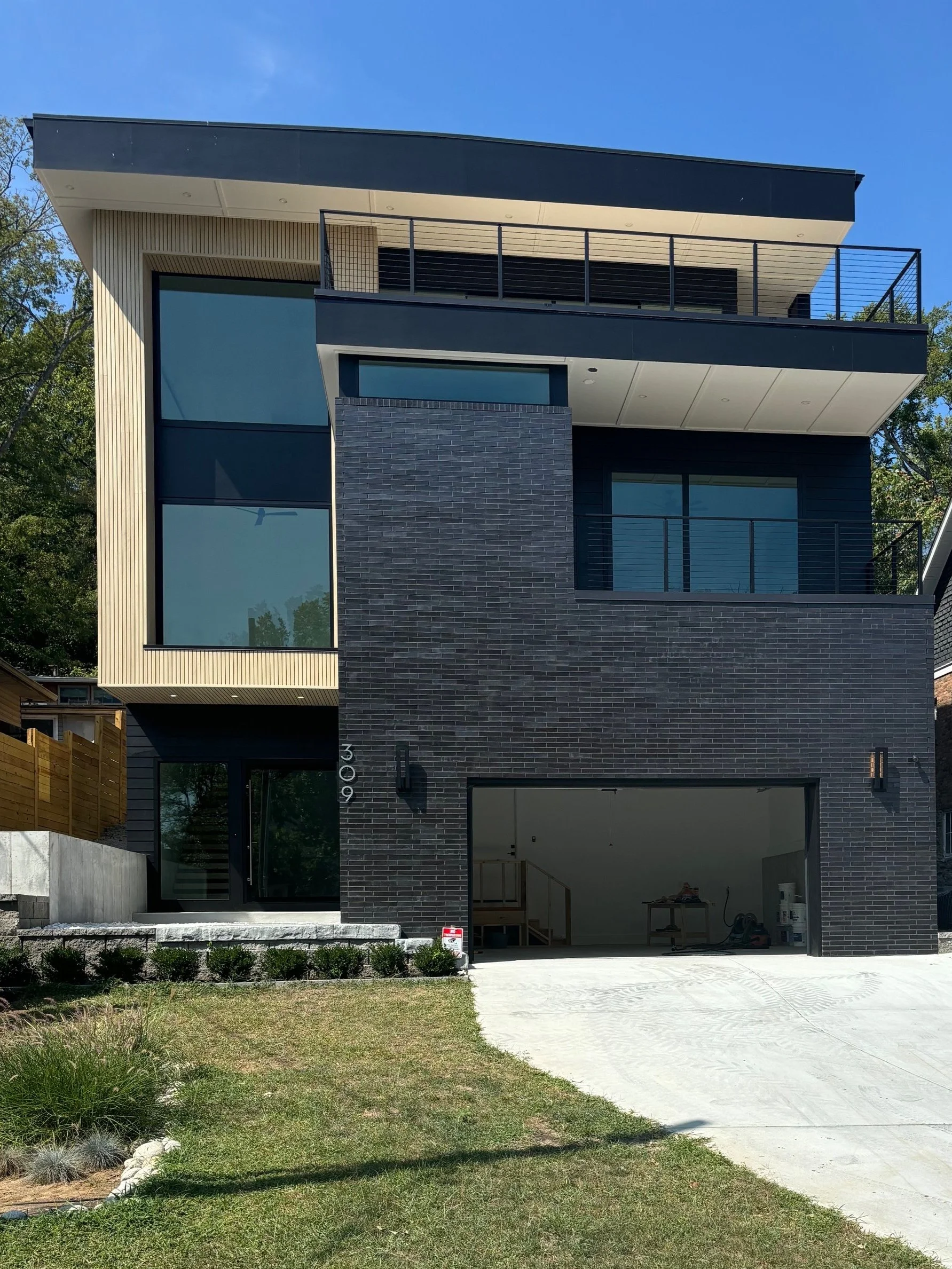 Modern multi-story house under construction with black brick facade, large windows, and a garage opening, surrounded by a grassy yard.
