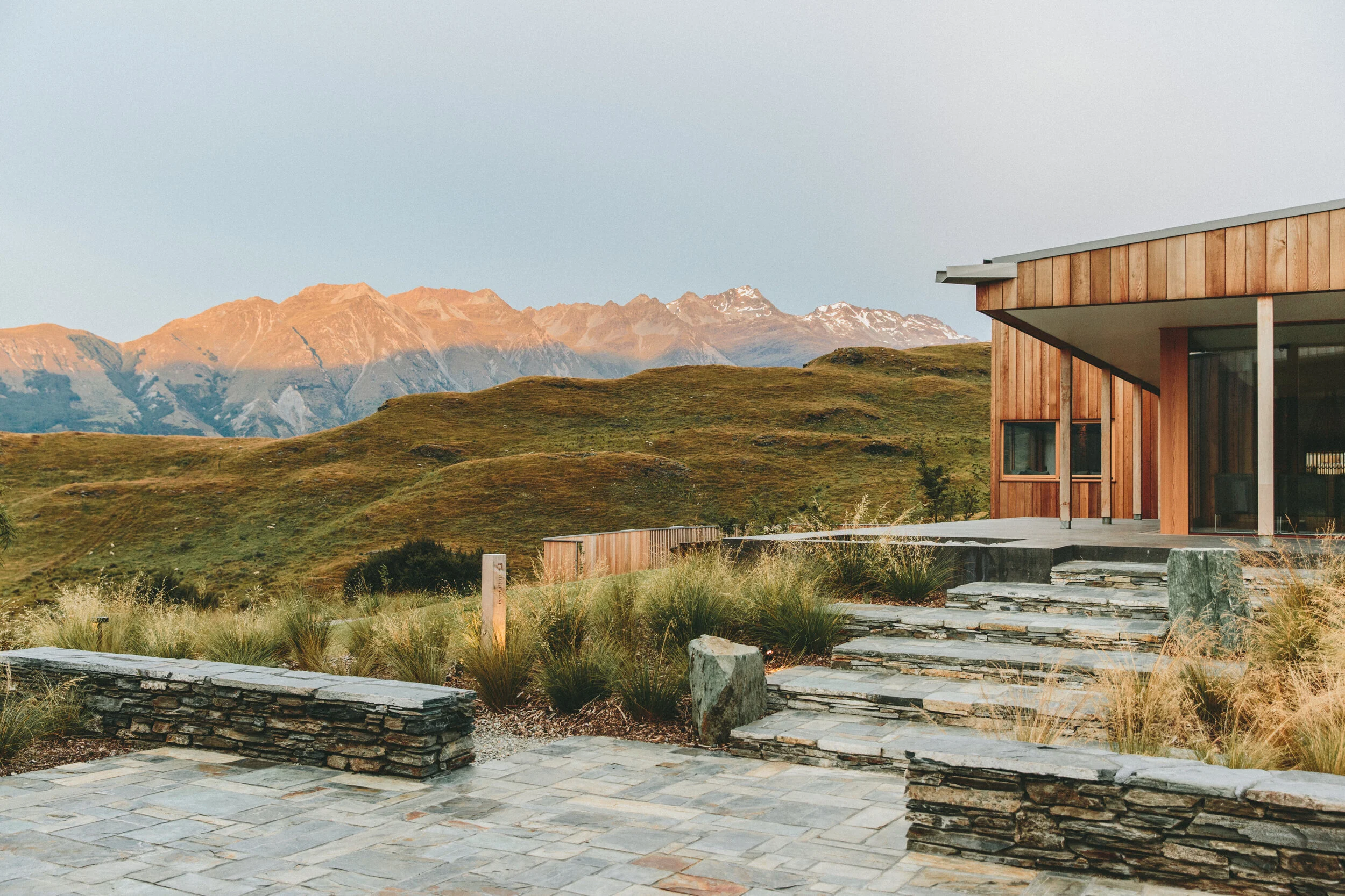 Modern wooden house with stone steps and patio, surrounded by green grass and bushes, mountains in the background at sunset.