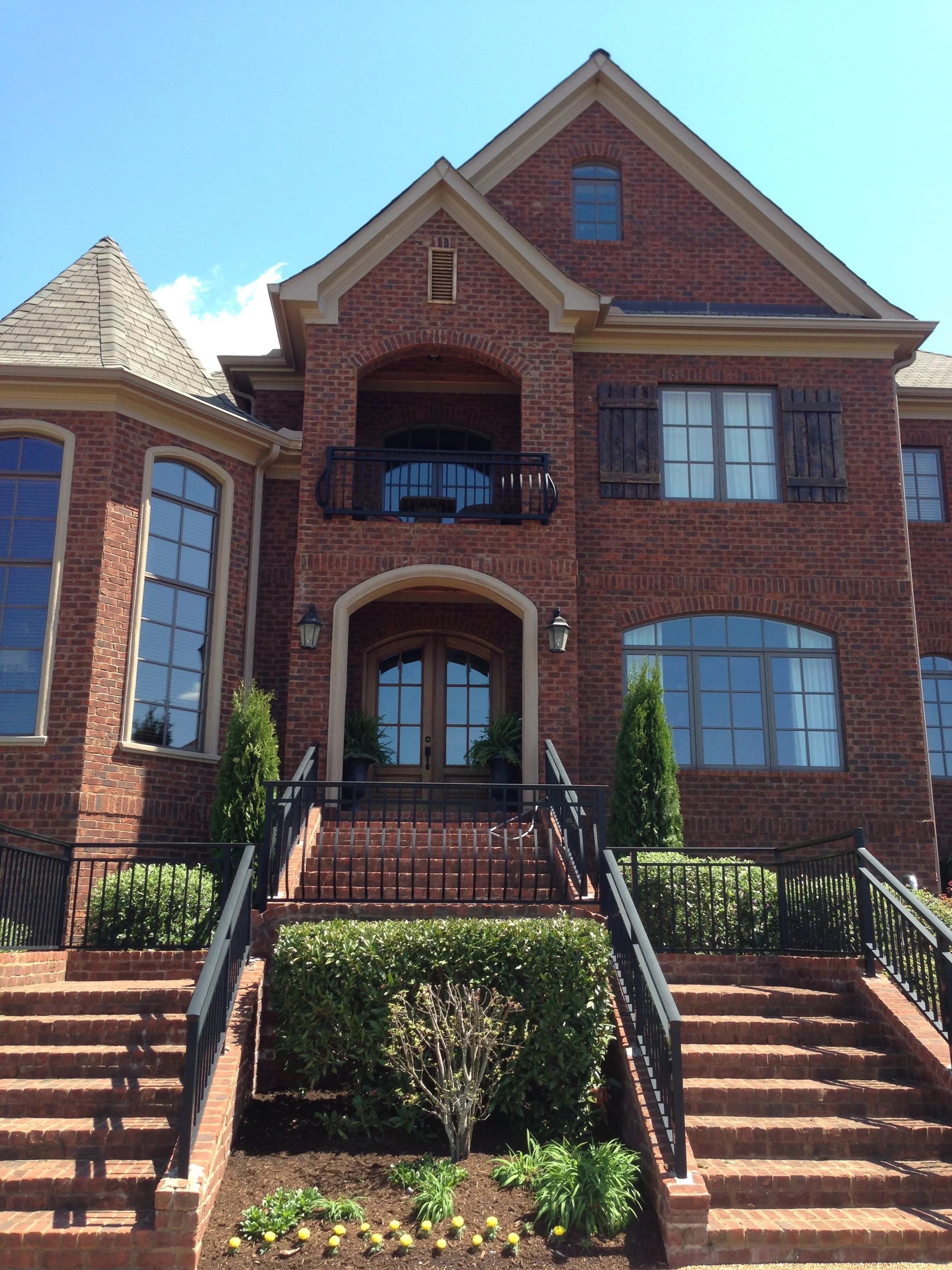 A large brick house with steps leading to a front door, surrounded by neatly trimmed bushes and plants, under a bright blue sky.