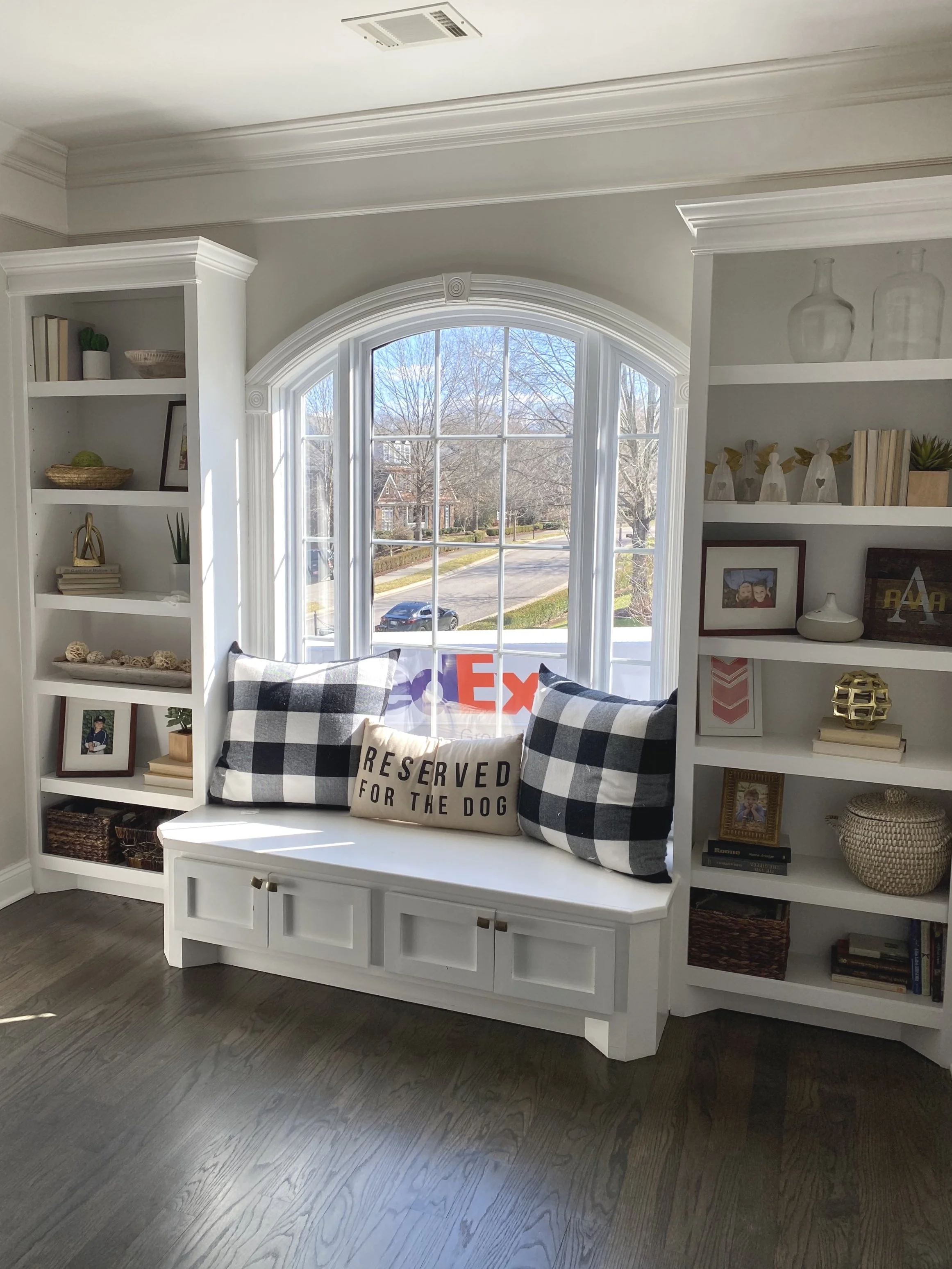 Living room window seat with black and white checkered pillows, a beige pillow that says "Reserved for the Dog," flanked by built-in white bookshelves filled with decor and books, over a hardwood floor.