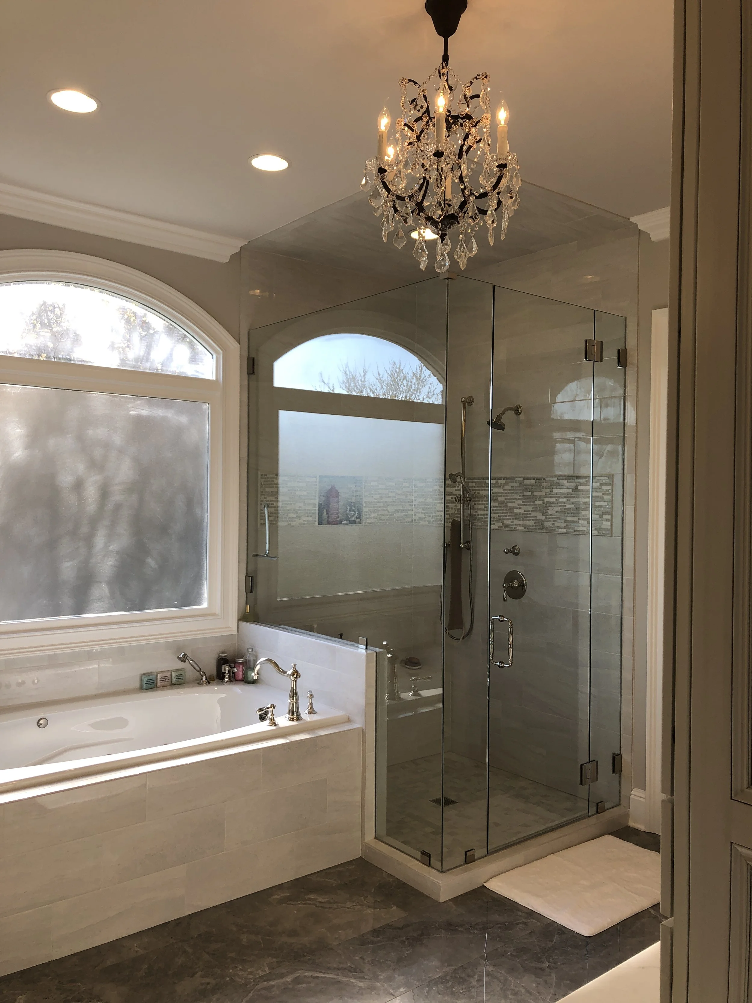 A bathroom with a bathtub, frosted window, glass shower enclosure, chandelier, and white rug.