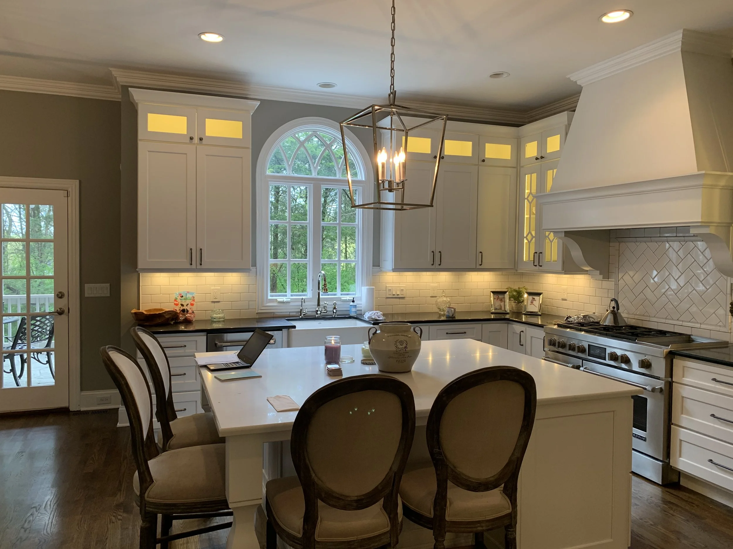 Bright kitchen with white cabinets, black countertops, and a central island. Large arched window with a view of greenery, white subway tile backsplash, and a stainless steel stove. Dining table with four chairs. Pendant light fixture. Door leading ou