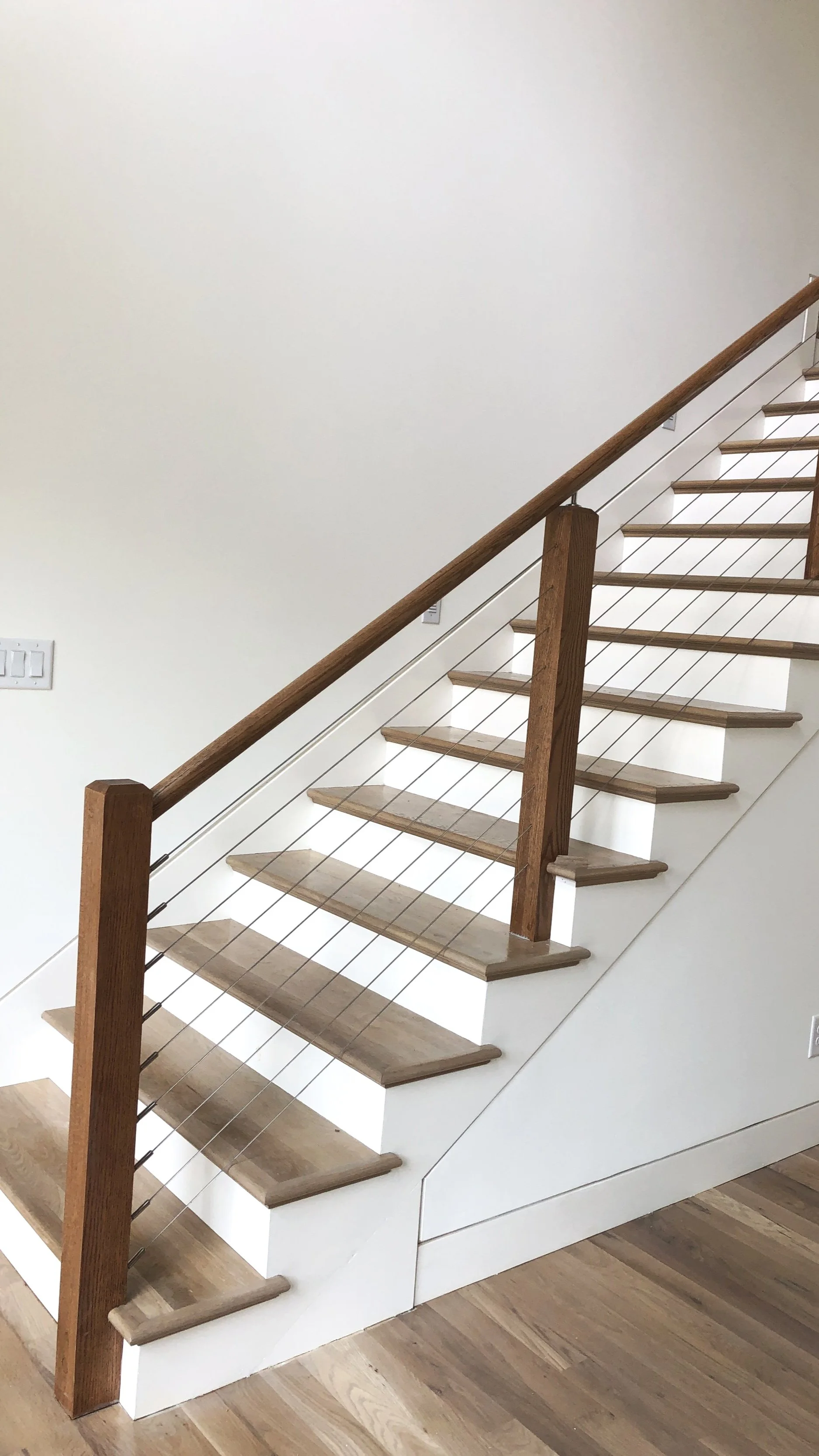 Wooden staircase with white risers, wooden treads, and a wooden handrail supported by metal cables, next to a white wall and light wood floor.