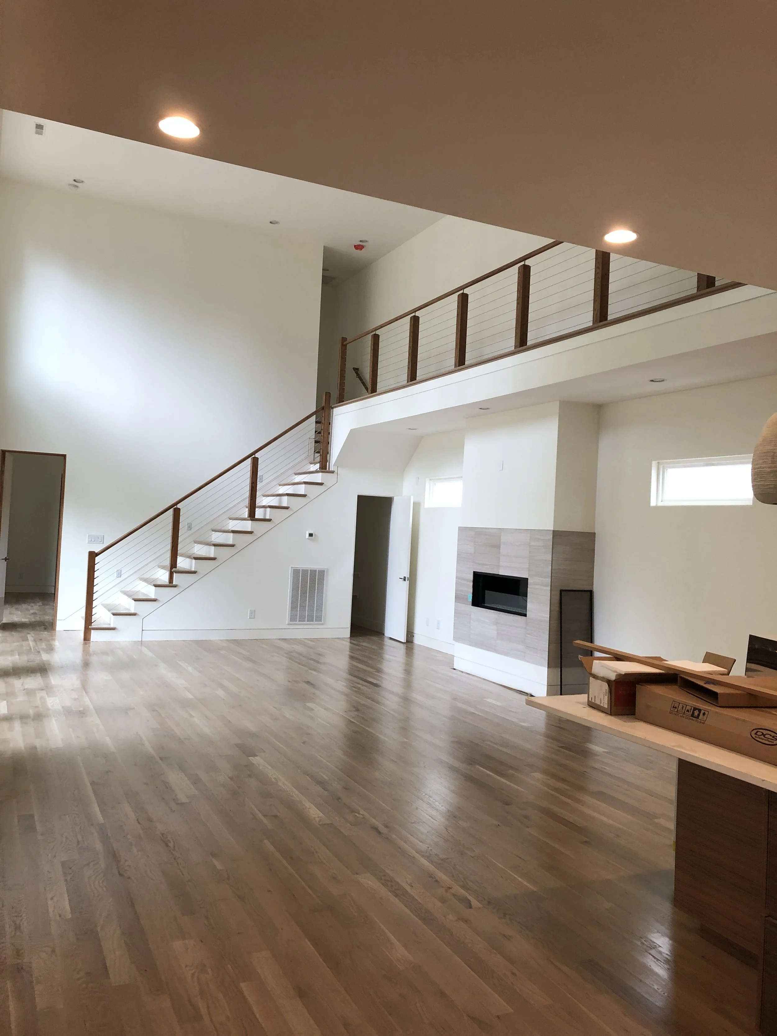 Empty living room with wooden floors, fireplace, staircase with wooden railing, and natural light from small windows.