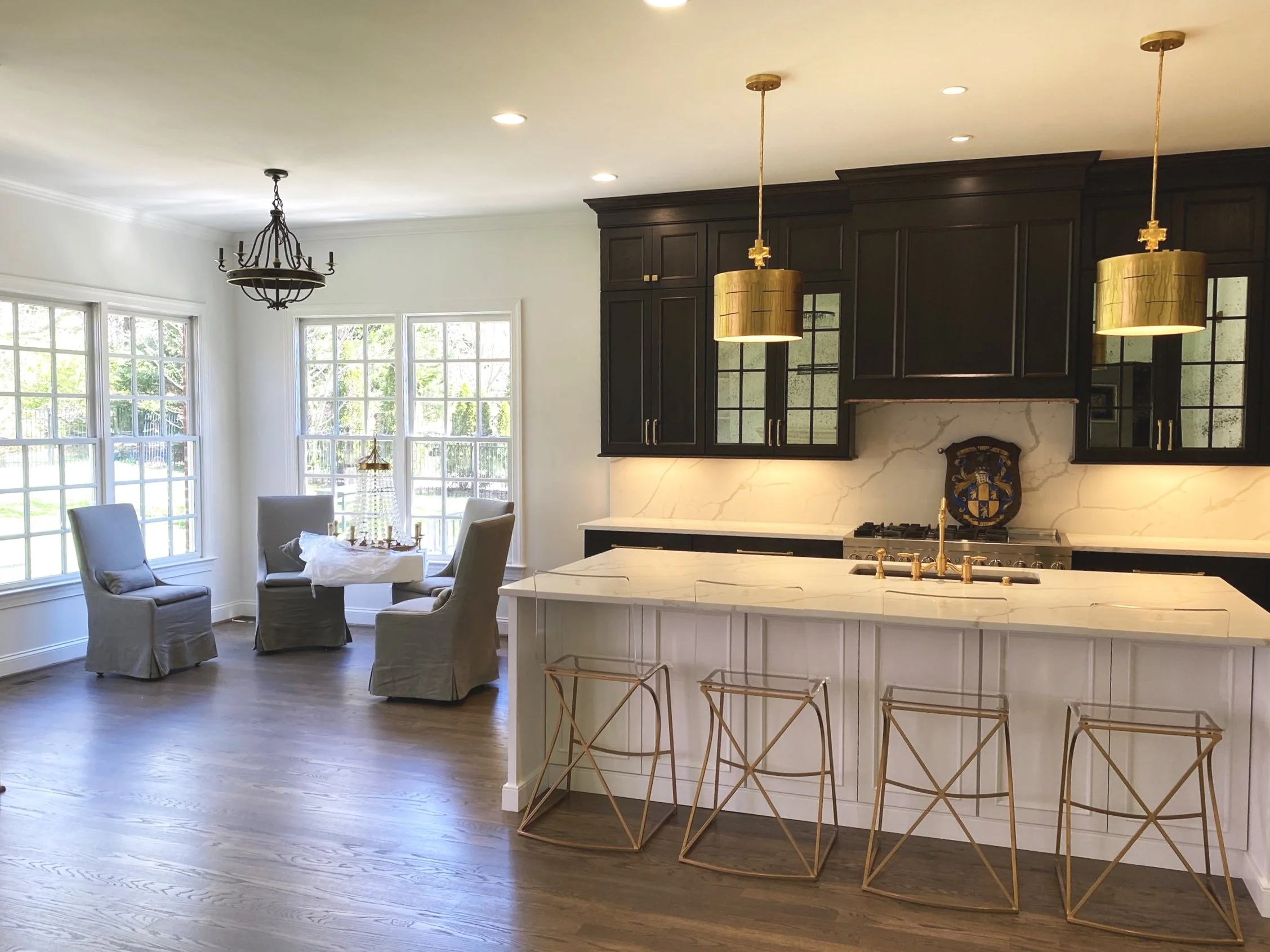 Bright kitchen and dining area with large windows, dark upper cabinets, white marble backsplash and counters, gold pendant lights, kitchen island with gold barstools, and a corner with a small round table and chairs.