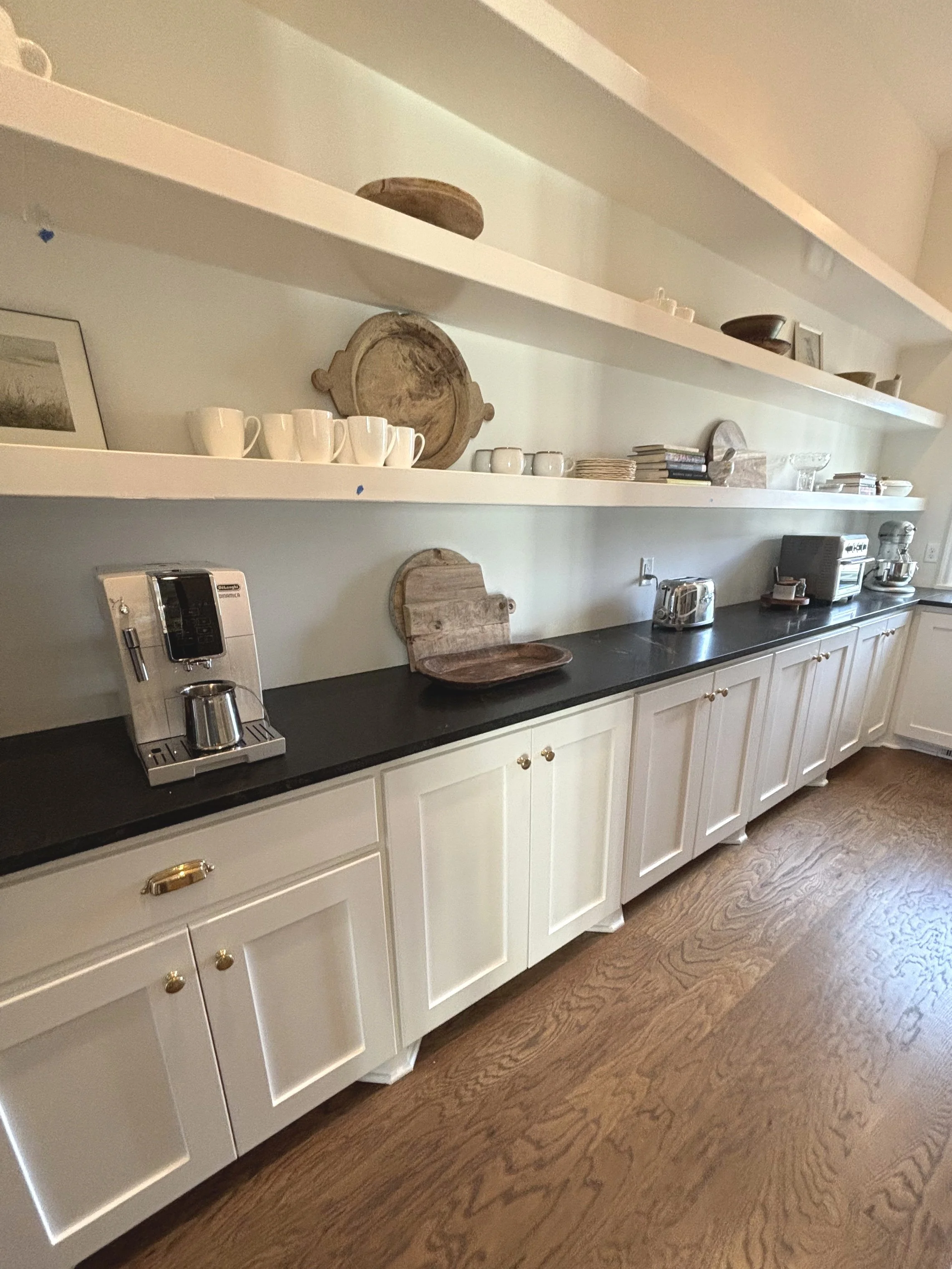 Kitchen counter with black countertop, white cabinets, open shelves with plates, cups, and decorative items, toaster, coffee machine, and stand mixer.