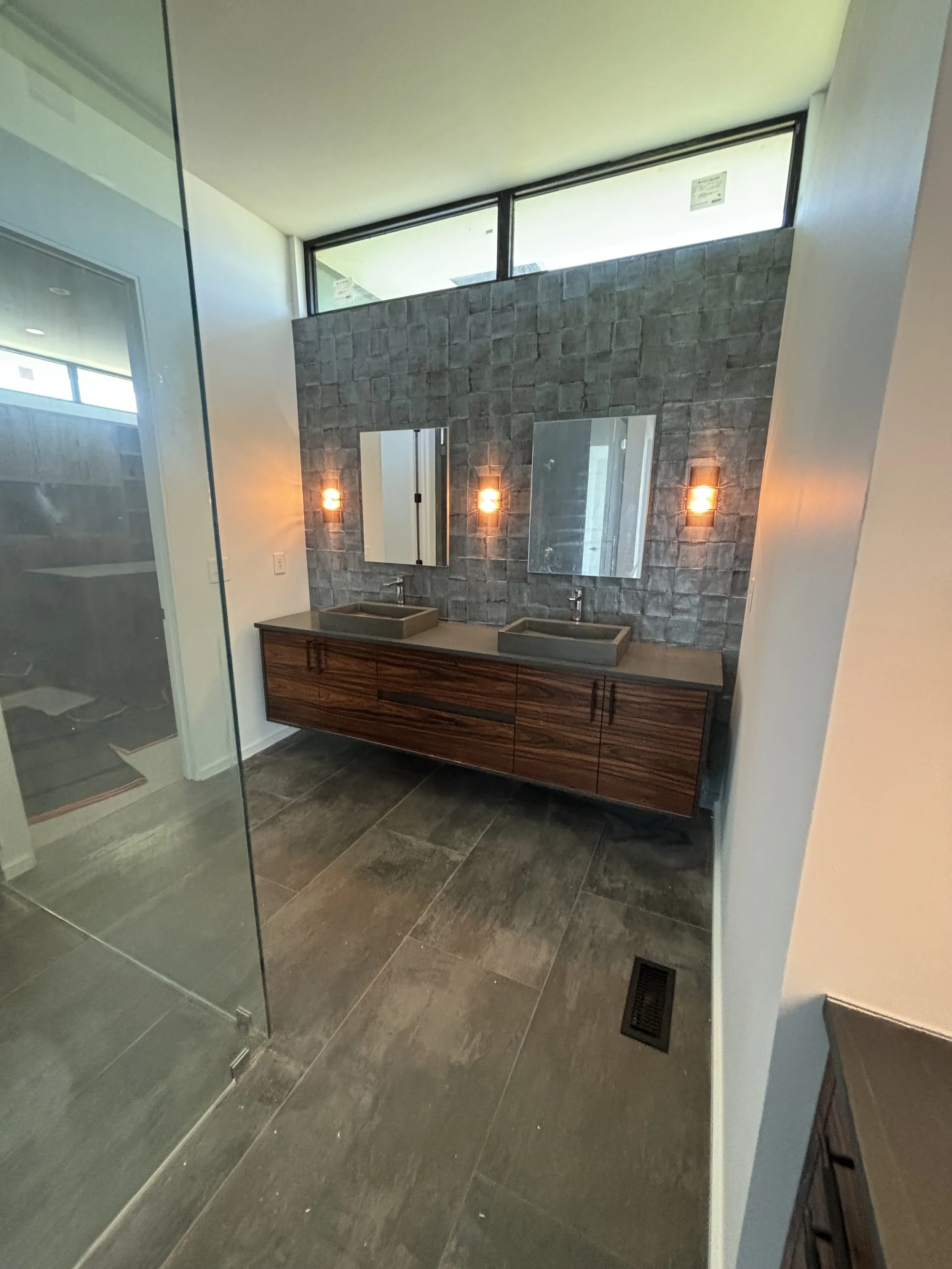Modern bathroom with gray tile flooring, dual sinks on a wooden vanity, gray textured tile wall with two mirrors and warm wall sconces, and a glass shower enclosure on the left.