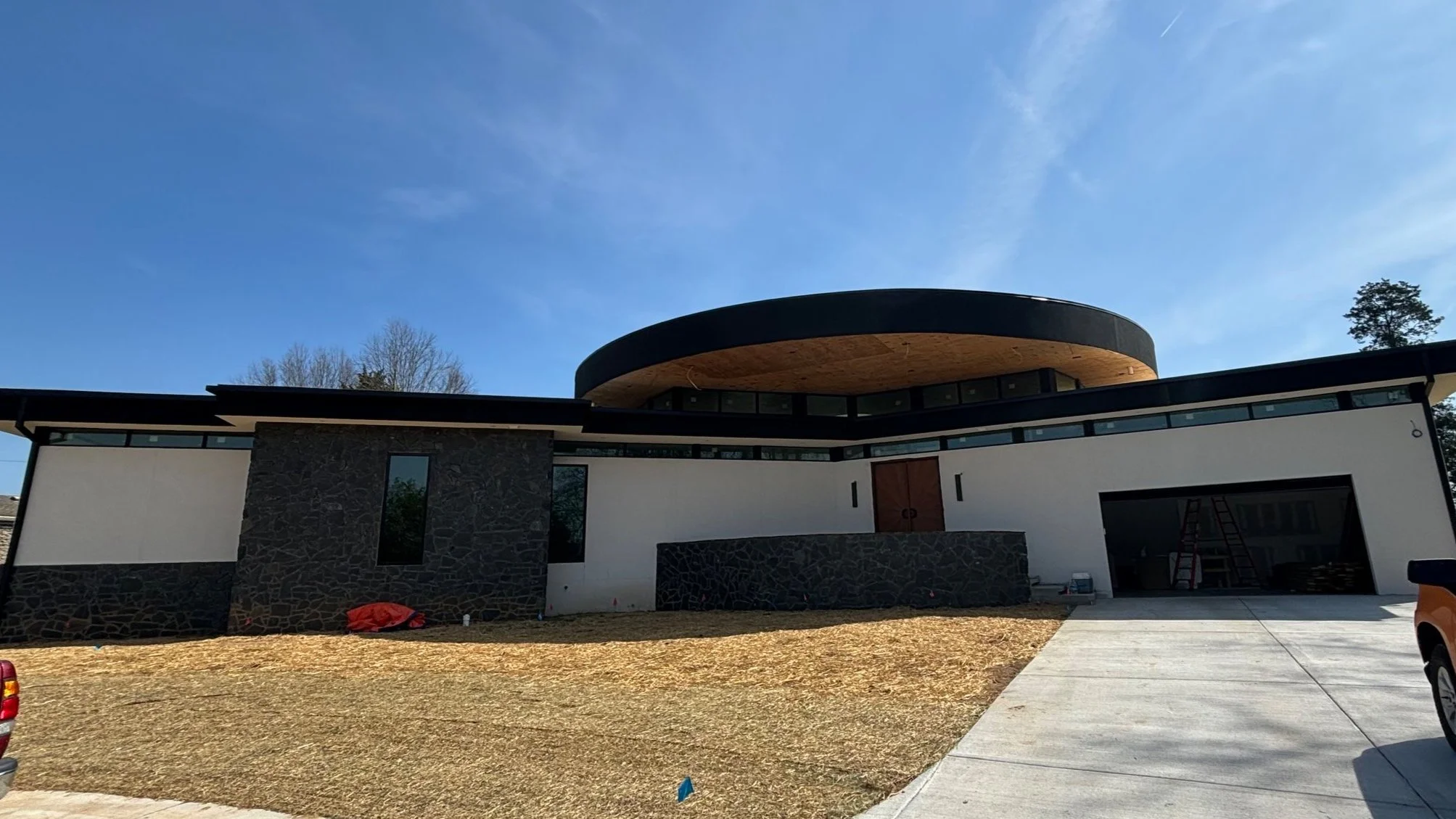 Modern house under construction with a round roof section, black and white exterior walls, and a garage, set against a bright blue sky.