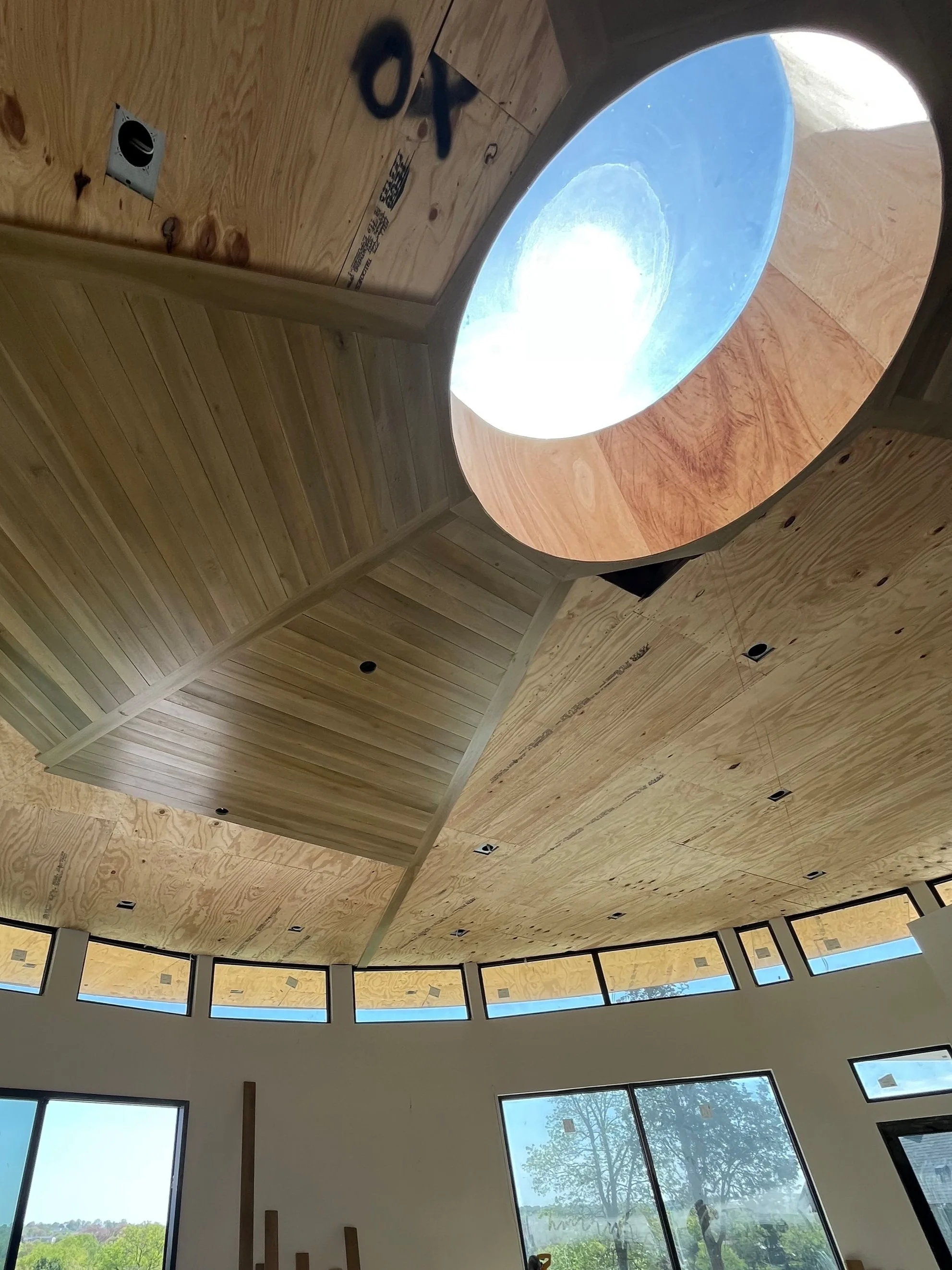 Interior view of a modern building with a round skylight and large windows, showcasing a wooden ceiling and green trees outside.