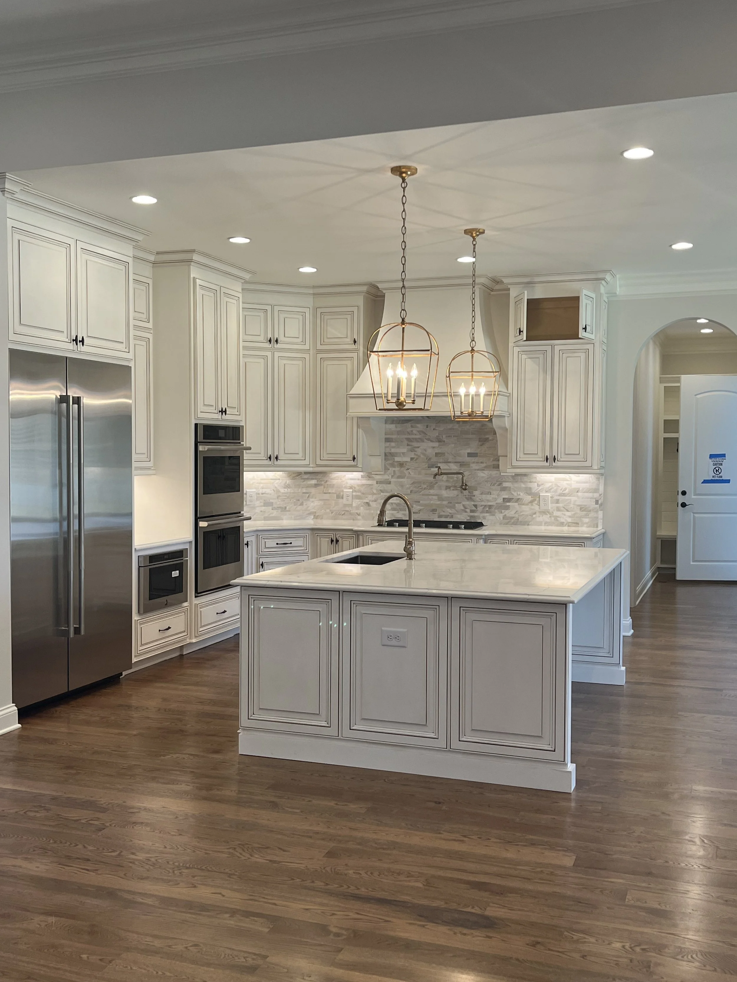Modern kitchen with white cabinetry, stainless steel appliances, and a large island with a marble countertop. Two pendant lights hang over the island, and recessed lighting is installed in the ceiling. The floor is hardwood.