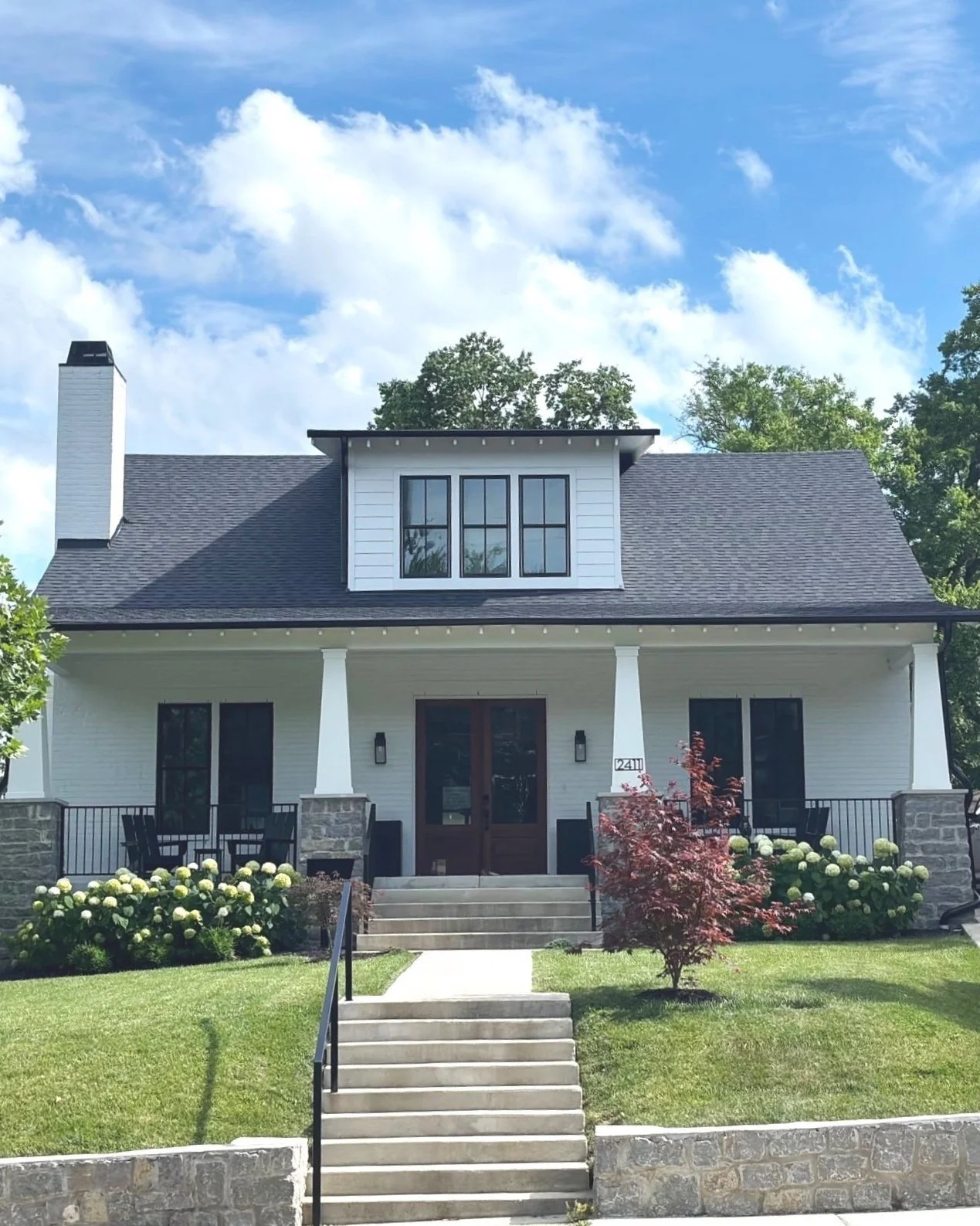 A white two-story house with a dark roof, set amidst green trees and a well-kept lawn, featuring front steps, a porch with black railing, and a dormer window on the upper level.