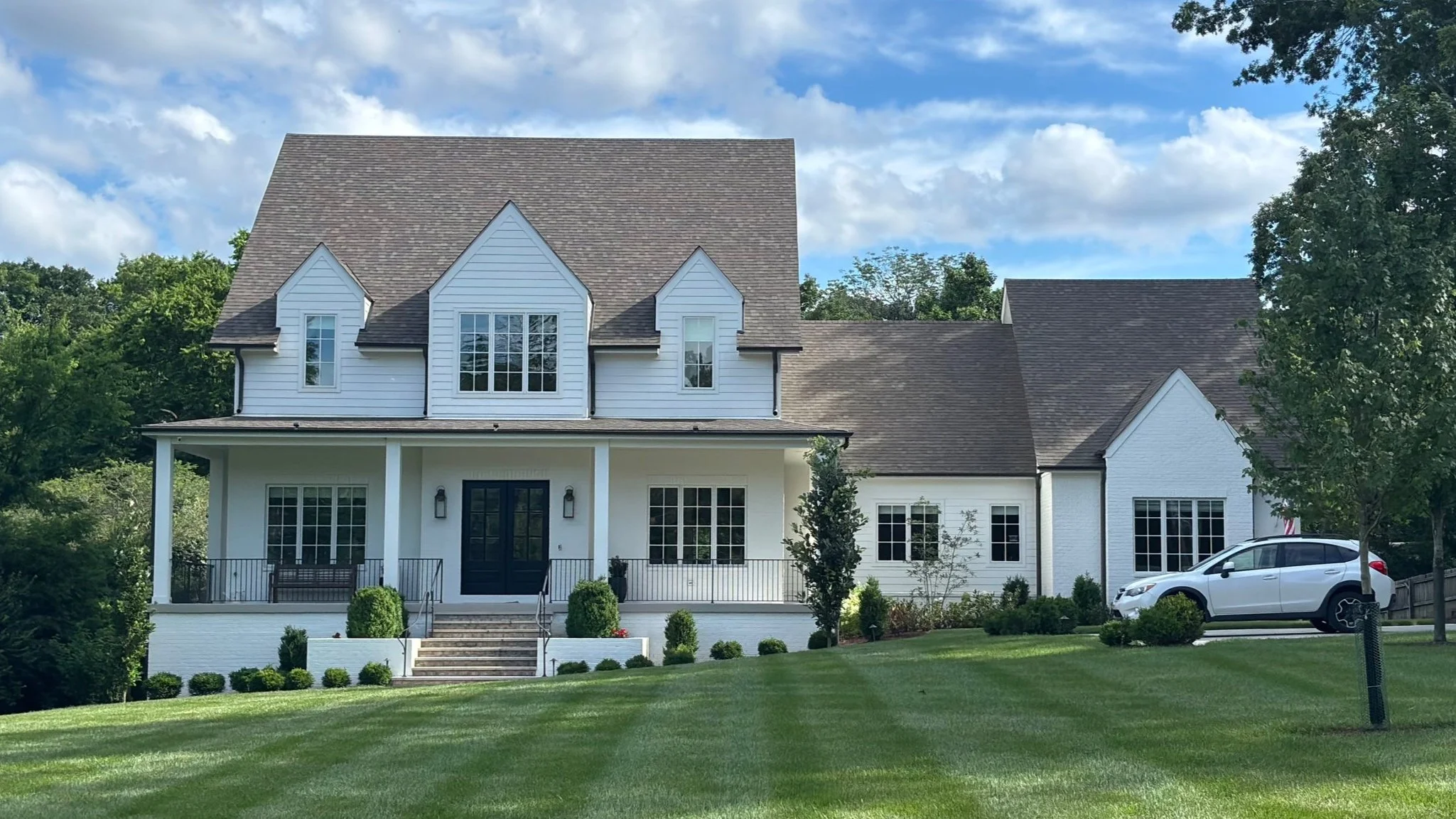 Large white house with black front door, multiple windows, three dormer windows on the roof, surrounded by green lawn, trees, and blue sky with white clouds.