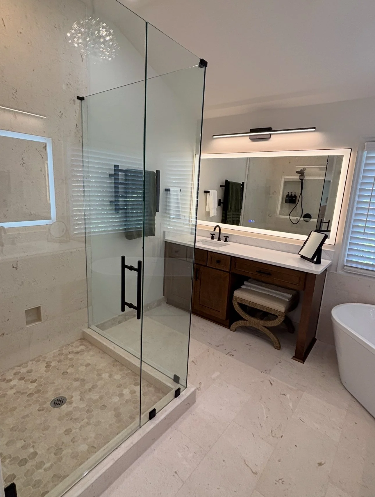 Modern bathroom with glass shower enclosure, wooden vanity with large mirror, white bathtub, and decorative tiles on the shower floor.
