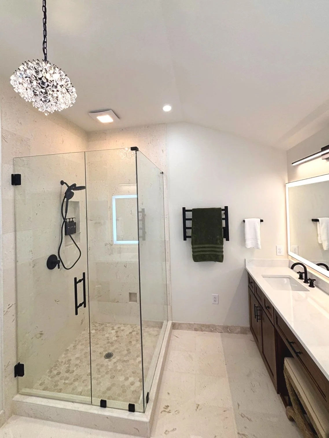 Modern bathroom with a glass shower enclosure, a white vanity with black fixtures, a wall-mounted towel rack with green and white towels, a large mirror with lighting, and a ceiling chandelier.