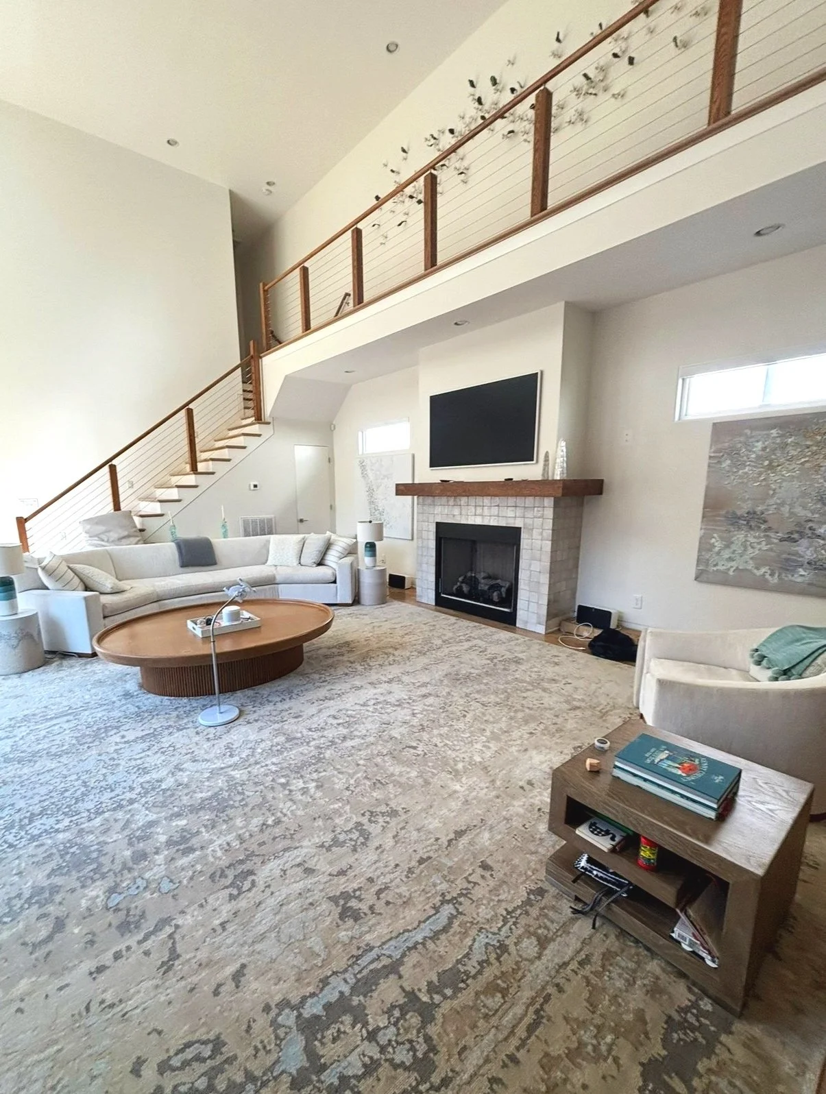 Living room with beige walls and a high ceiling, large off-white sectional sofa, round wooden coffee table, fireplace with tiled surround and a flat-screen TV above, area rug, side tables with lamps, and artwork on the wall. There is a staircase with