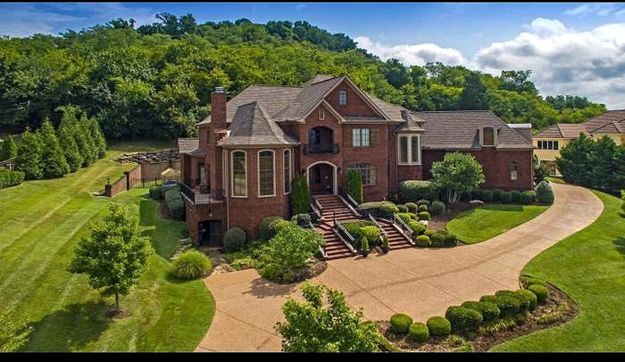 Large brick house with a curved driveway, surrounded by green lawn and landscaped bushes, with a wooded hill in the background.