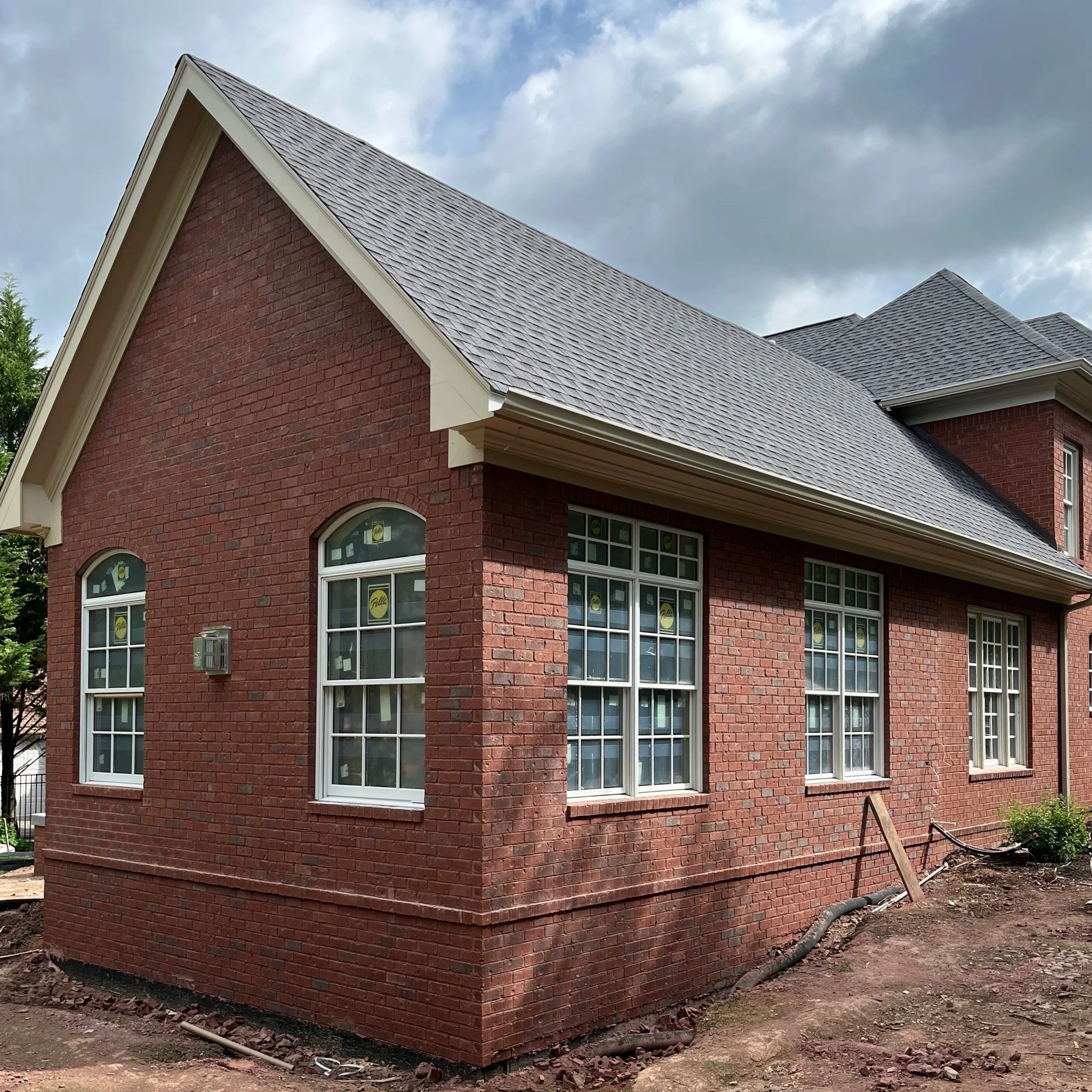 A brick house with multiple white-framed windows, a sloped gray shingle roof, and a small garden area near the right corner, under a cloudy sky.