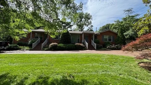 A row of three brick houses with front stairs surrounded by trees and bushes, with a green lawn in the foreground during daytime.
