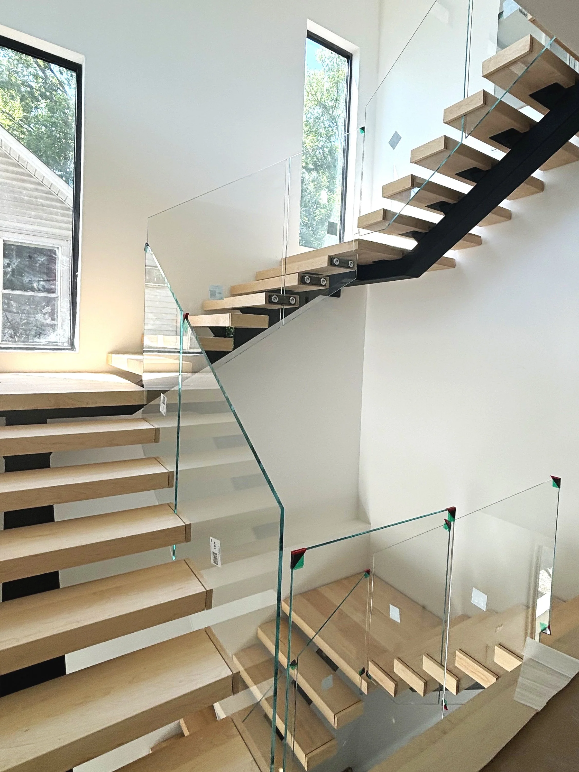 Modern staircase with wooden steps, black supports, and glass railings leading to an upper floor in a bright room with large windows.