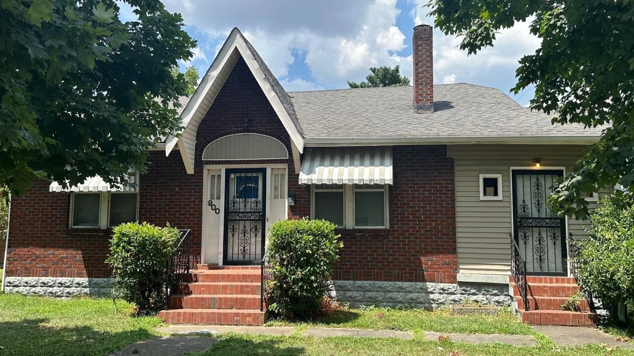 A house with brick and siding exterior, two front doors with black wrought iron security doors, brick stairs leading to each door, surrounded by green bushes and trees, under a cloudy sky.