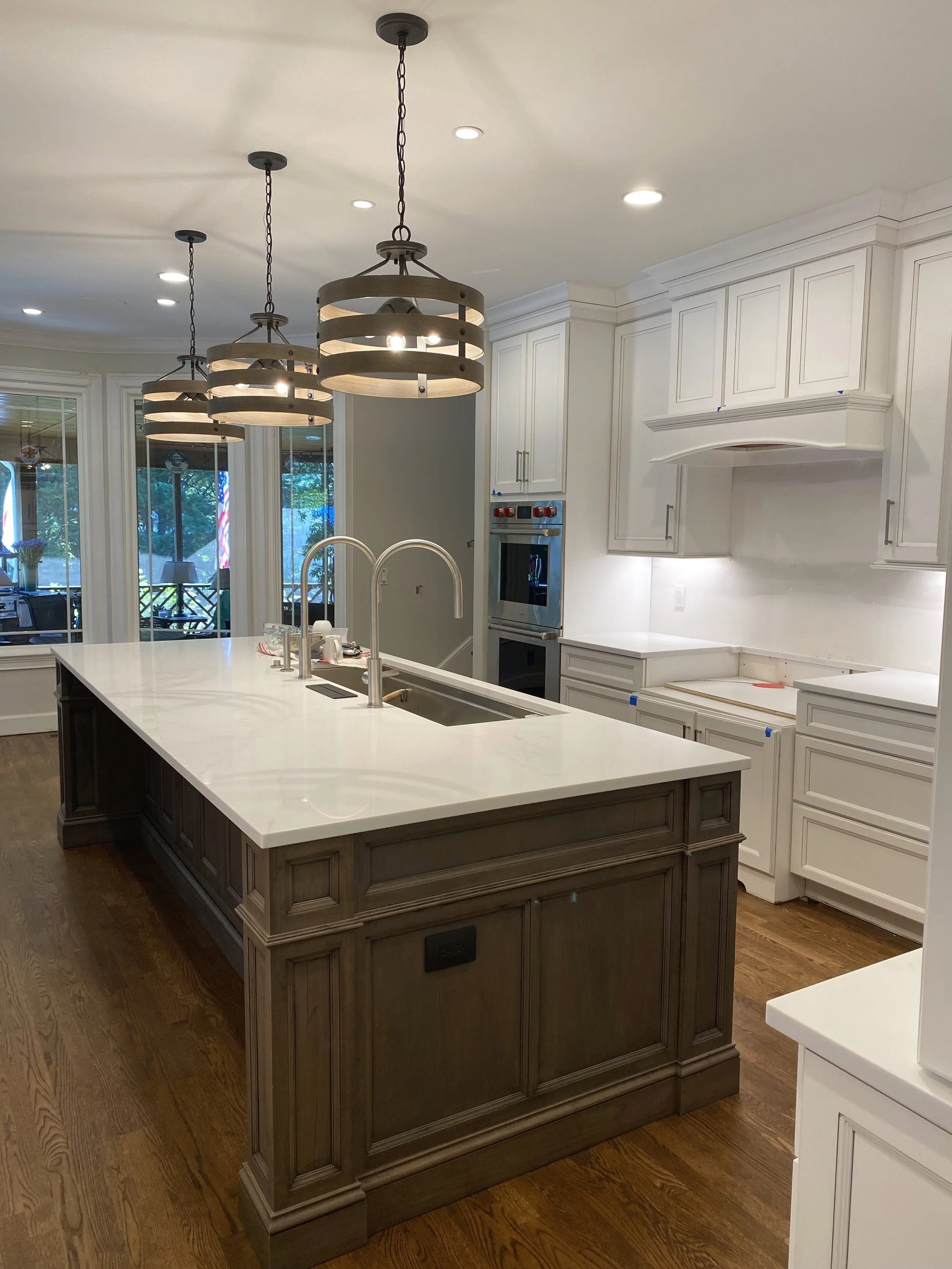 Modern kitchen with a large island, white cabinets, and wooden flooring. Overhead lighting fixtures hang above the island, and a view of the outdoors is visible through windows.