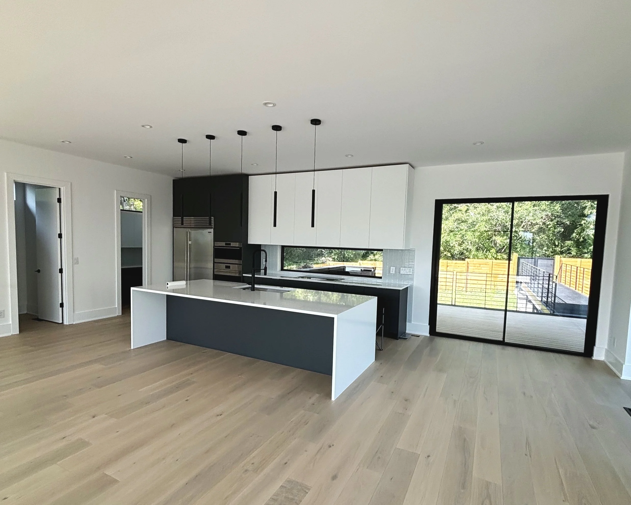 Modern kitchen with black and white cabinets, a large island with a white countertop, pendant lighting, large sliding glass door leading to outdoor deck, and light wood flooring.