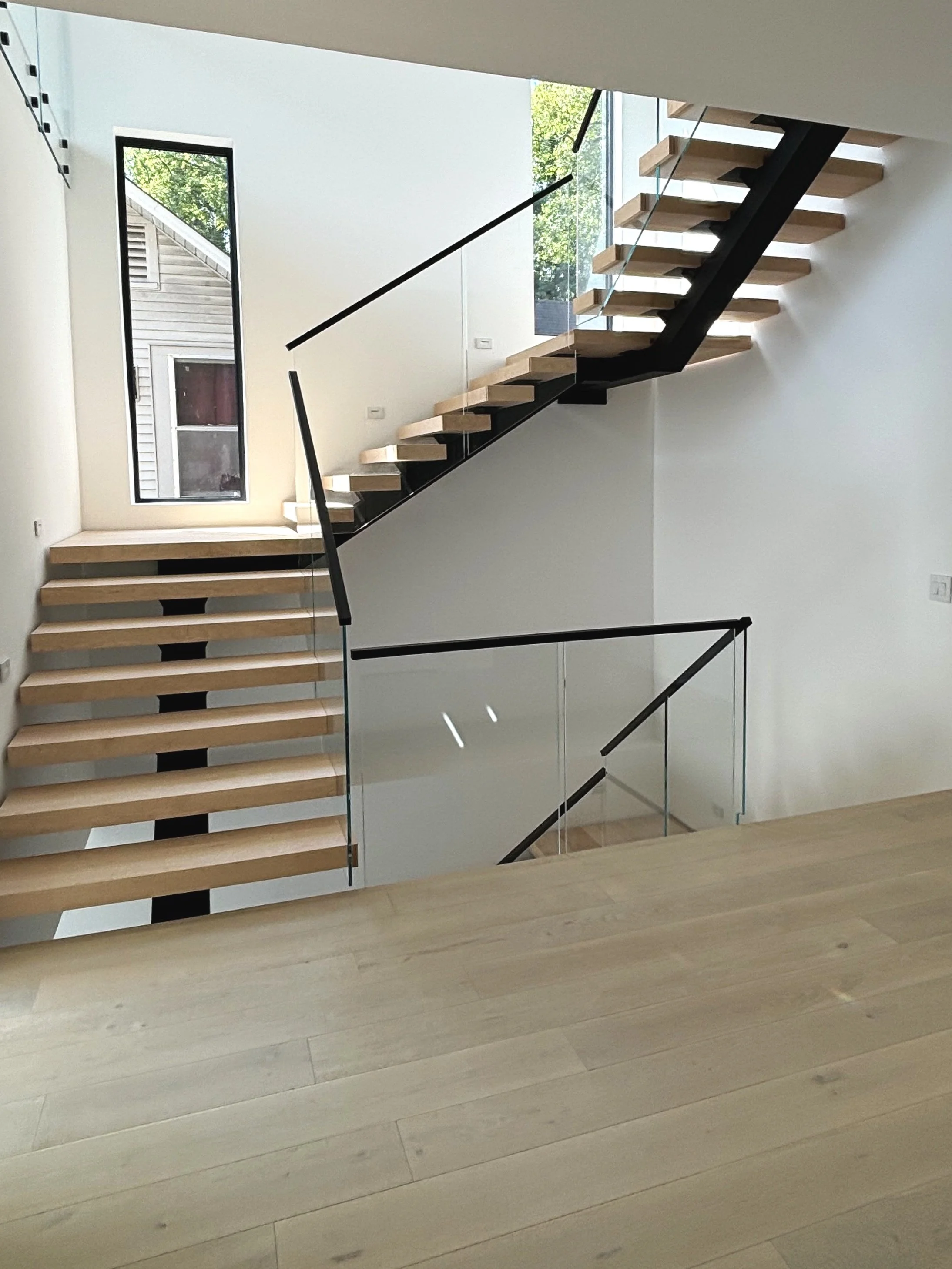 Interior view of a modern staircase with wooden steps, black supports, and a glass railing inside a bright, white-walled house, with a window providing natural light.