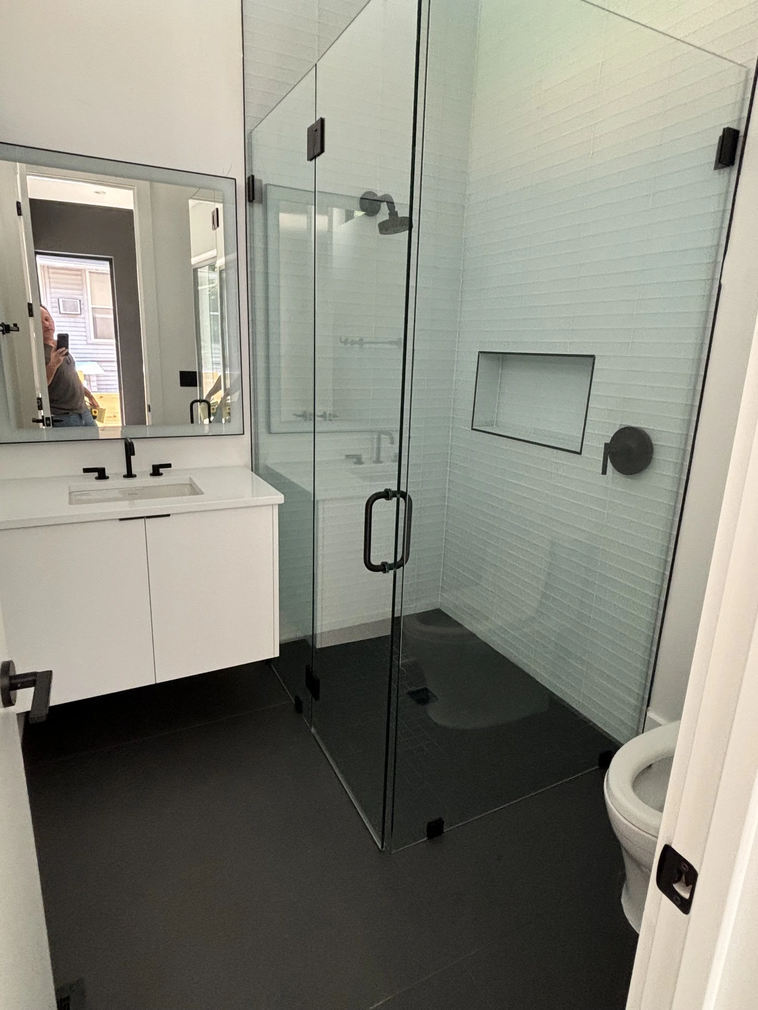 Modern bathroom with a glass shower enclosure, a white vanity with a mirror, and a toilet. The shower has white tiled walls with a built-in shelf and black fixtures.