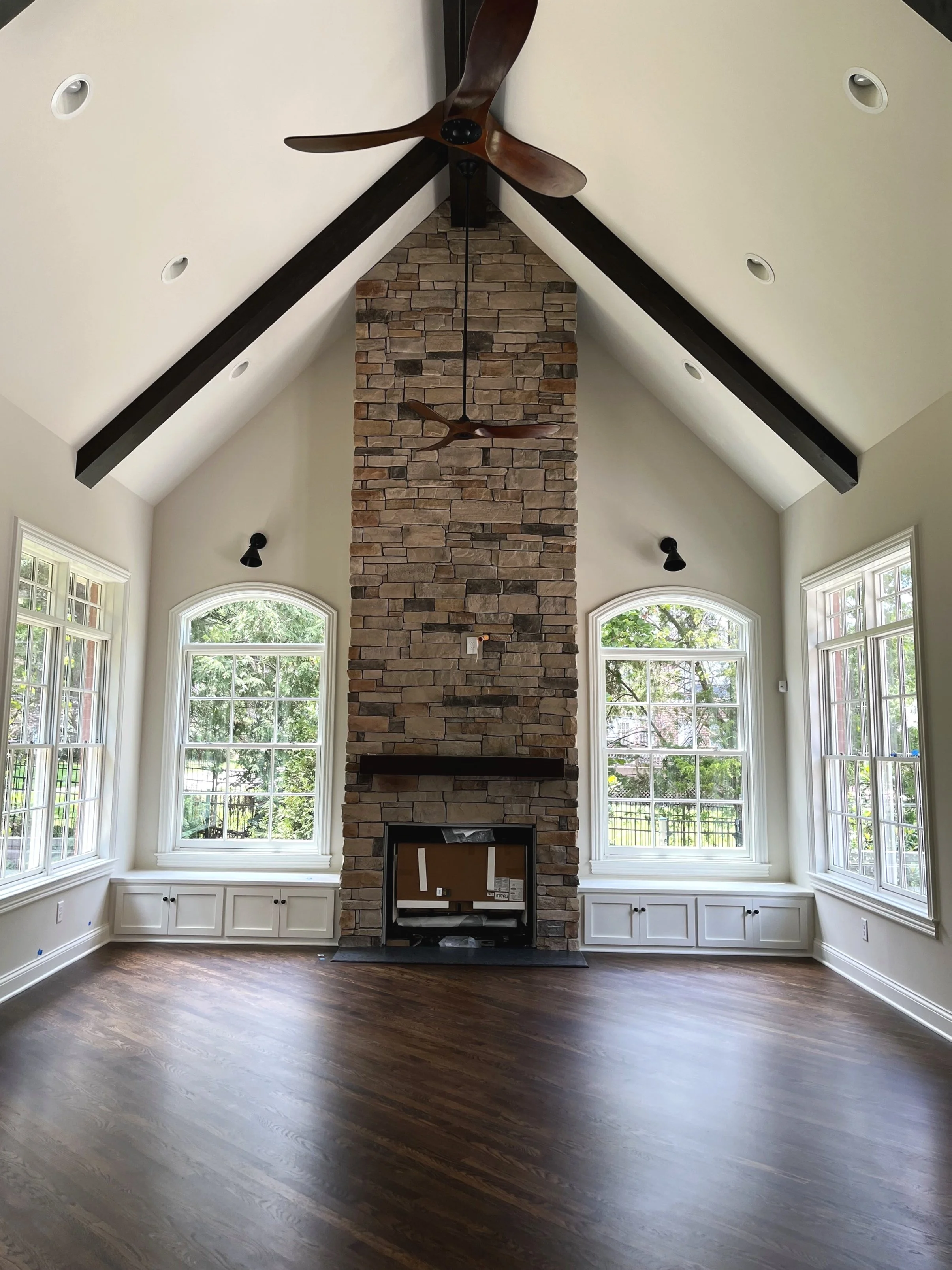 Empty living room with vaulted ceiling, stone fireplace, large windows, wooden floors, and ceiling fan.
