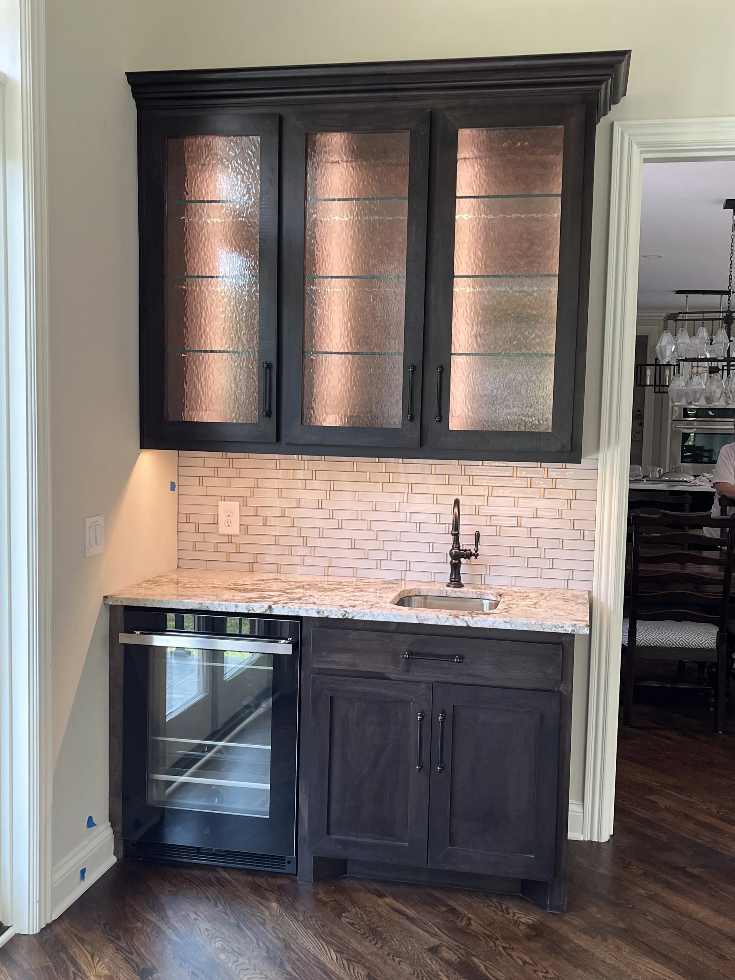 Miniature kitchen with dark wood cabinets, granite countertop, small sink, wine fridge, brick backsplash, and hardwood floors.