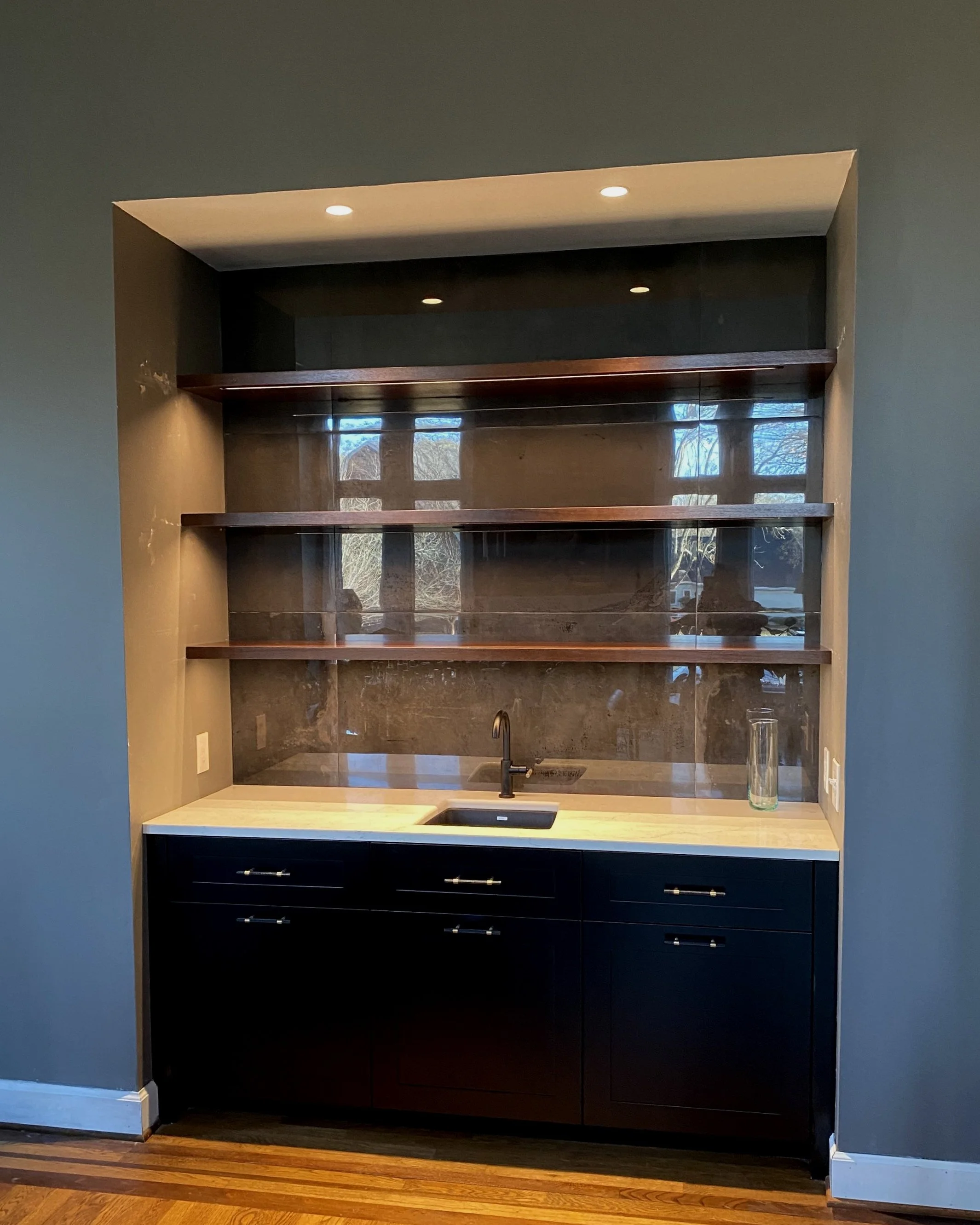 Modern kitchen nook with black cabinets, a white marble countertop, a black sink with a single faucet, three dark wooden open shelves above, grey walls, and a window reflecting trees outside.