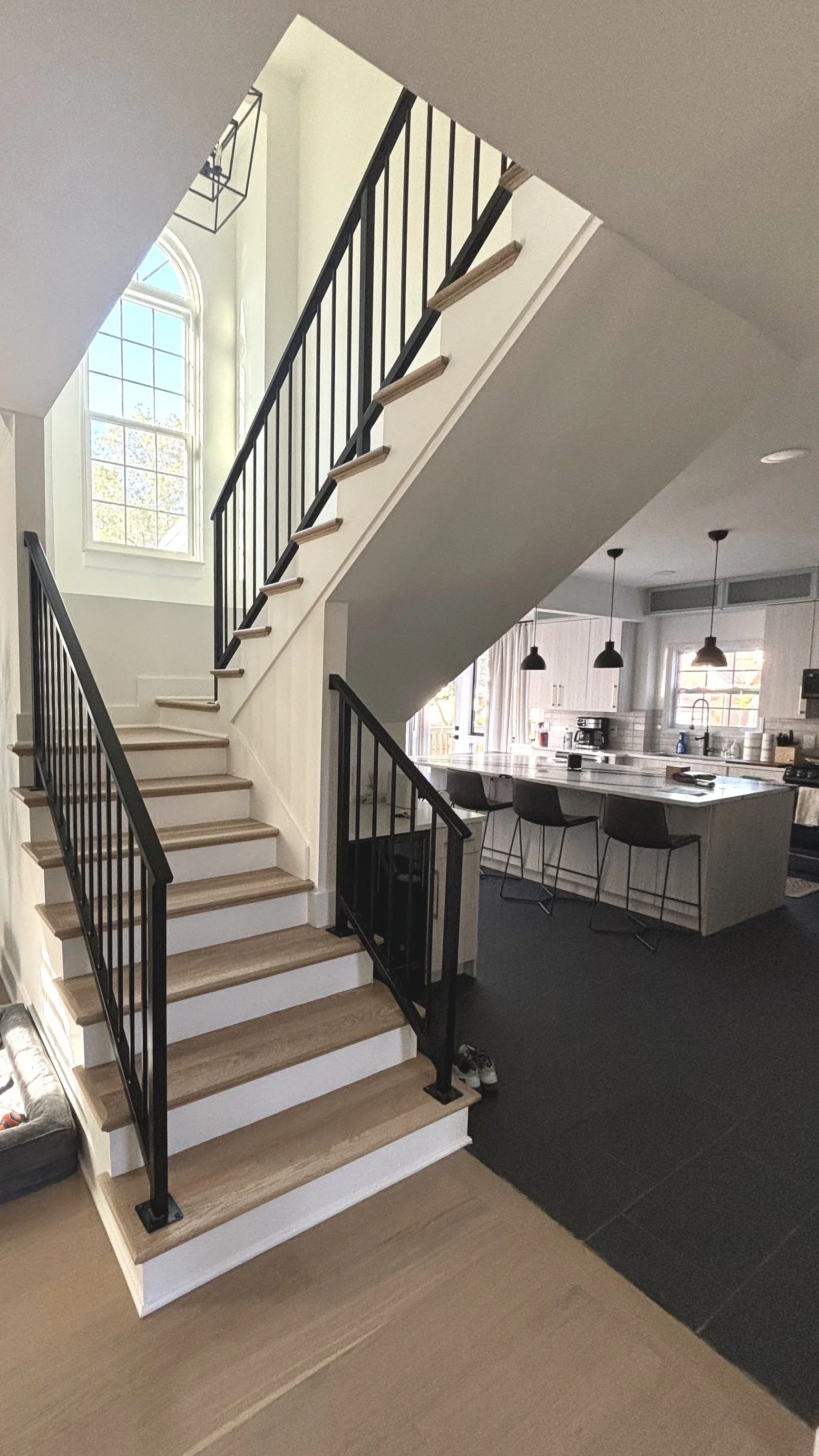 Inside view of a modern home showing a staircase with wooden steps and black metal railing, leading to an upper floor. Behind the staircase is a large window with an arched top. In the background, a kitchen with a large island, black bar stools, pend