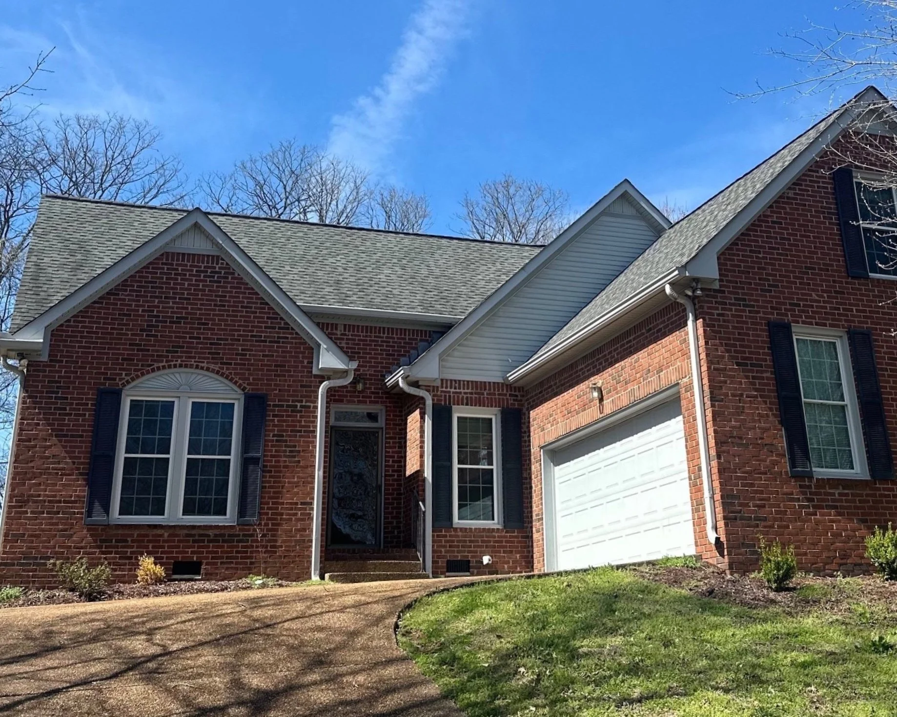 A two-story red brick house with a white garage door, black shutters, and a front pathway leading to the entrance, surrounded by a lawn and leafless trees under a blue sky.