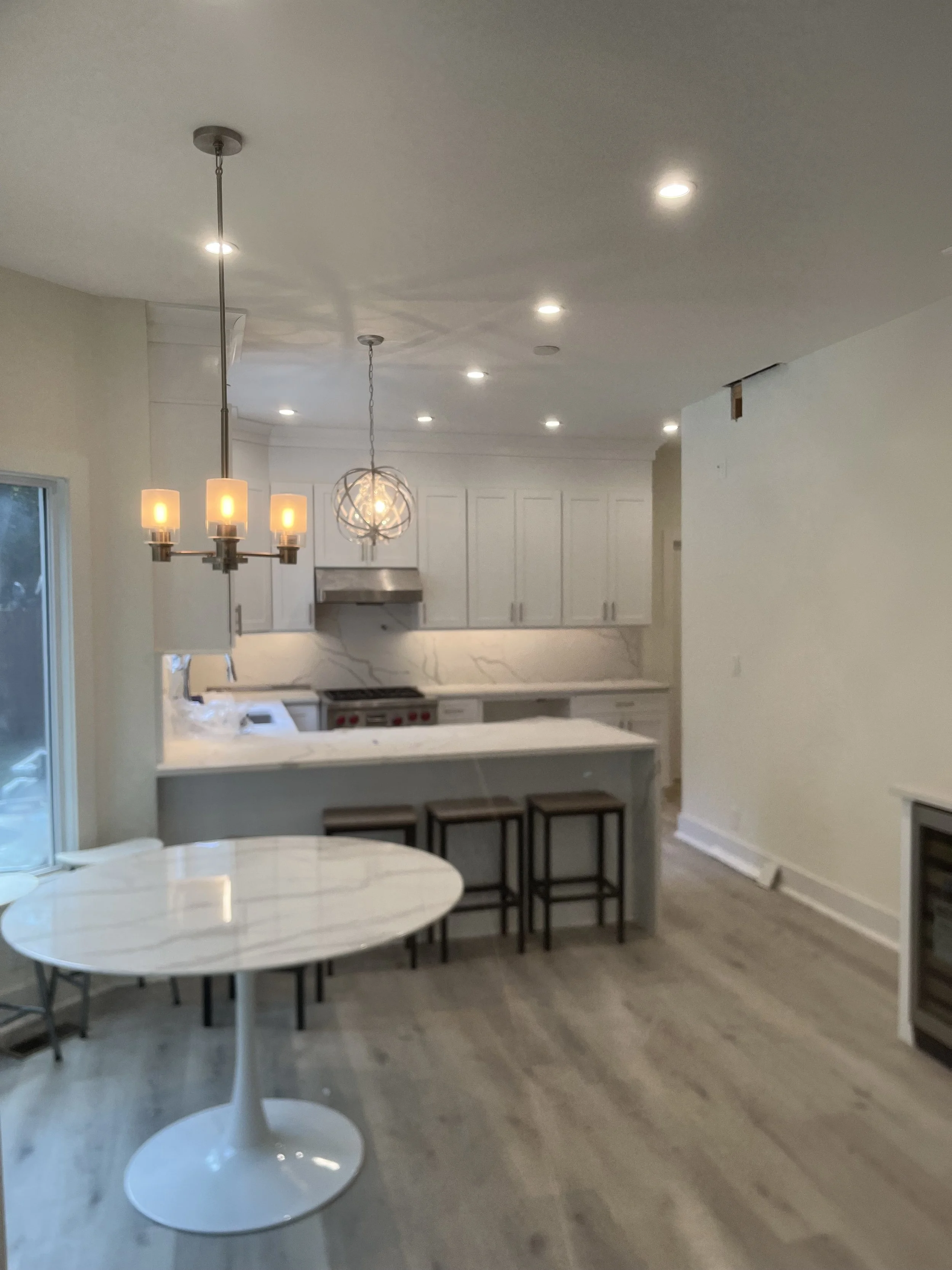 Open-concept kitchen and dining area with white cabinets, marble countertops, stainless steel appliances, and a round white marble table with three chairs. Recessed lighting and hanging light fixtures over the table and kitchen island. Wooden flooring and a sliding glass door on the left.