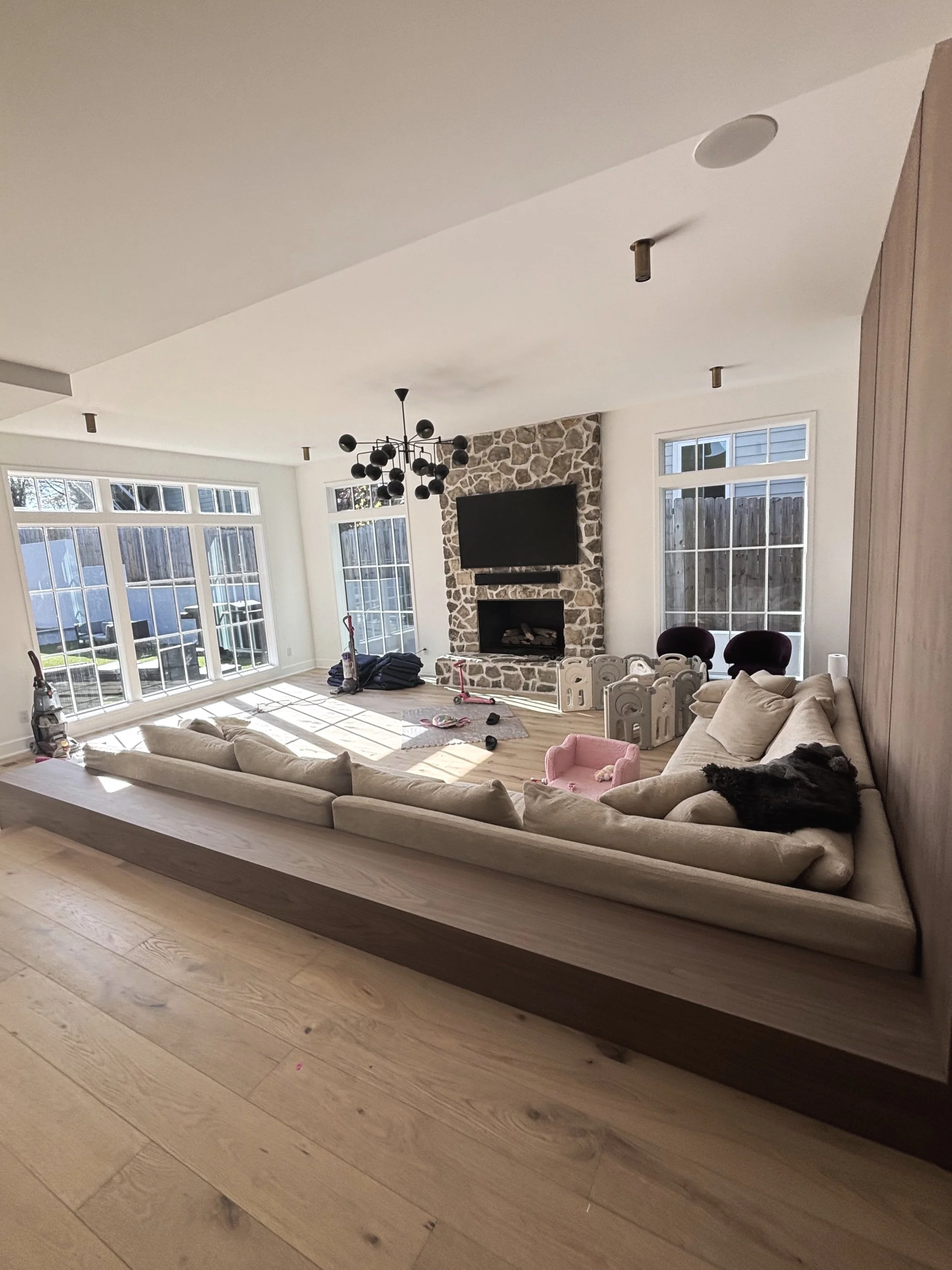 Living room with large off-white sectional sofa, stone fireplace with mounted TV, modern black chandelier, hardwood flooring, and large windows letting in natural light.
