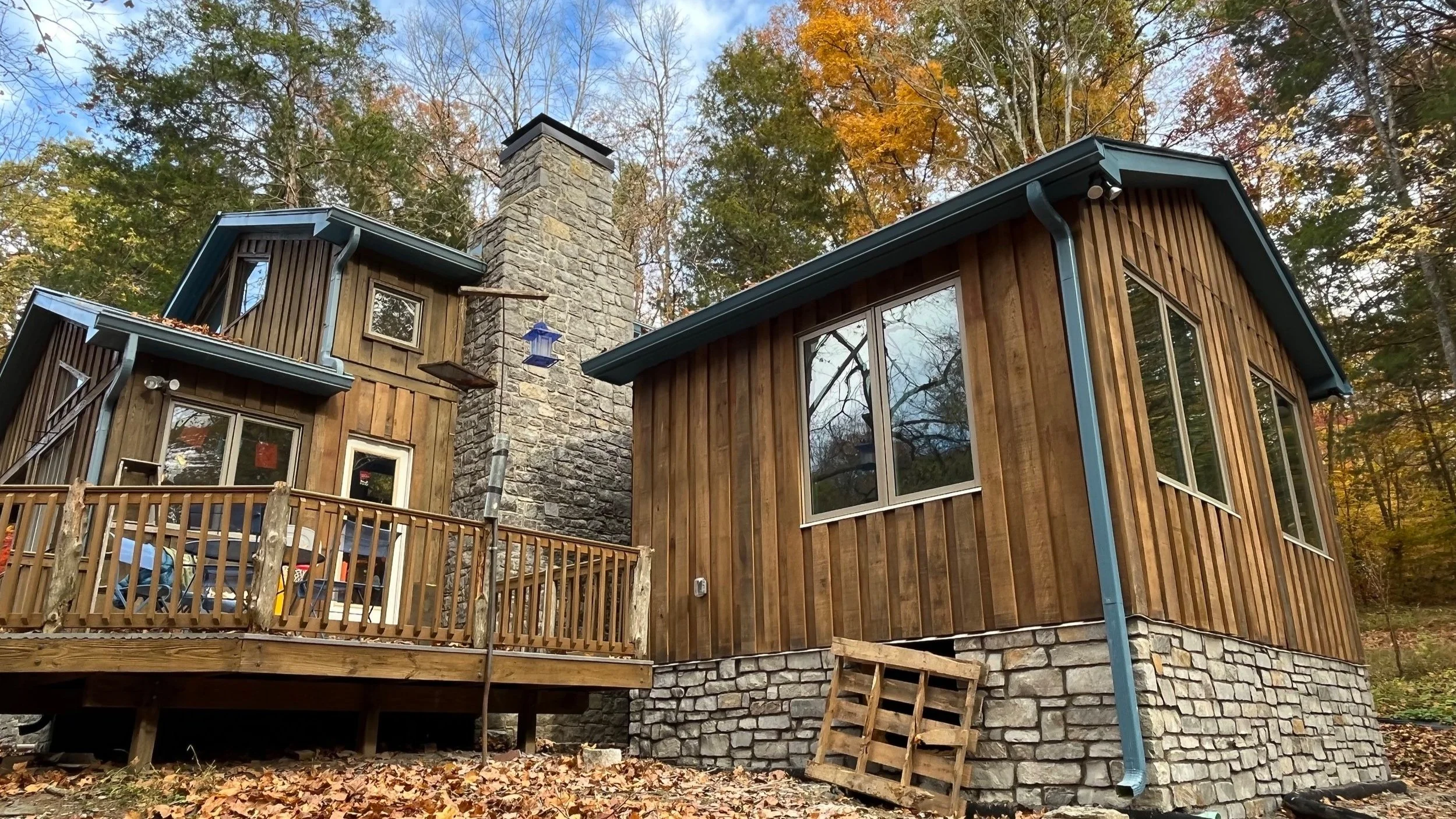 A house with wooden siding, large windows, and a stone chimney, surrounded by autumn trees and fallen leaves.
