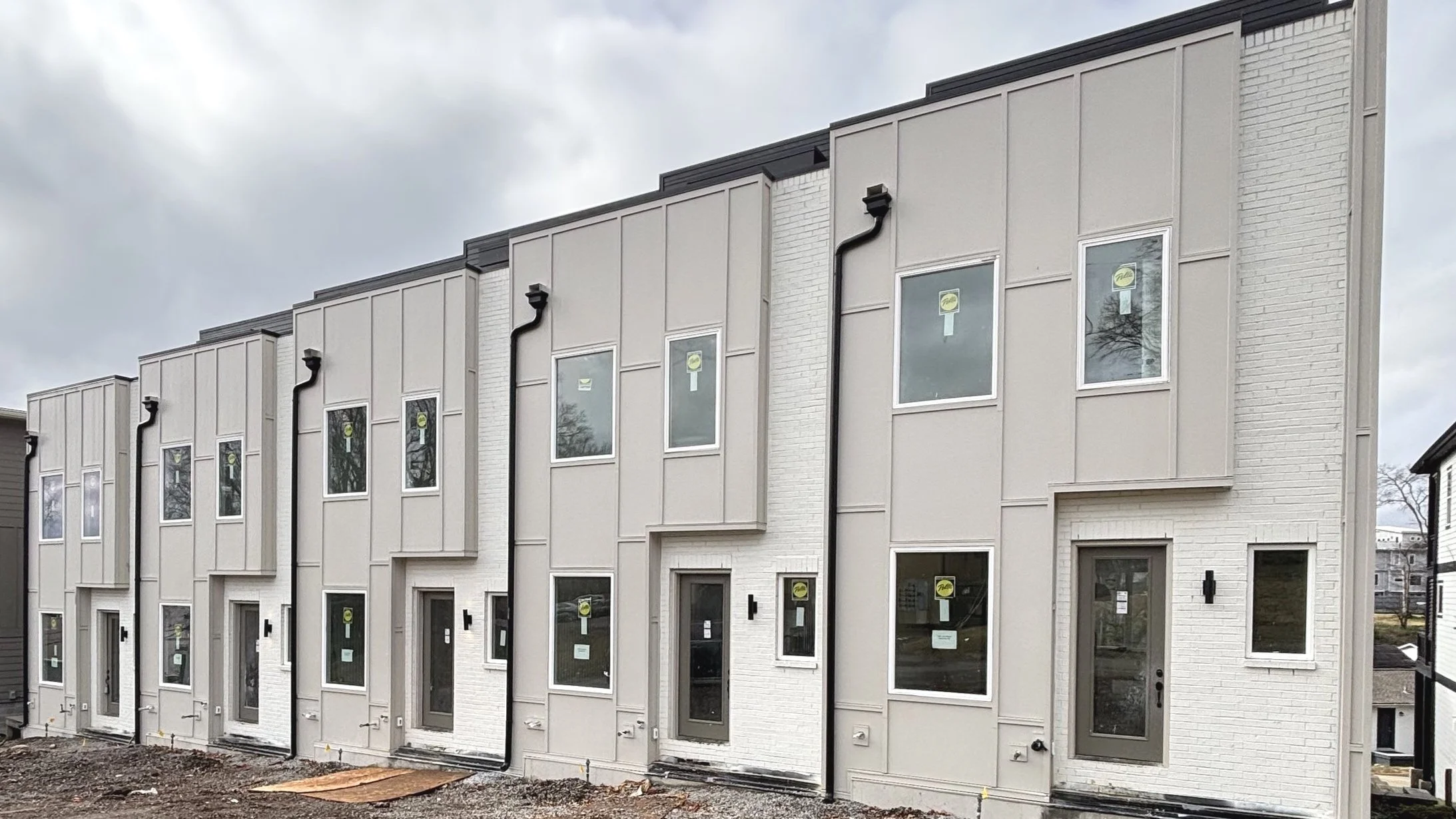 Newly constructed multi-story townhouses with modern white and gray exterior finishings, multiple windows, and black exterior lighting fixtures, under a cloudy sky.