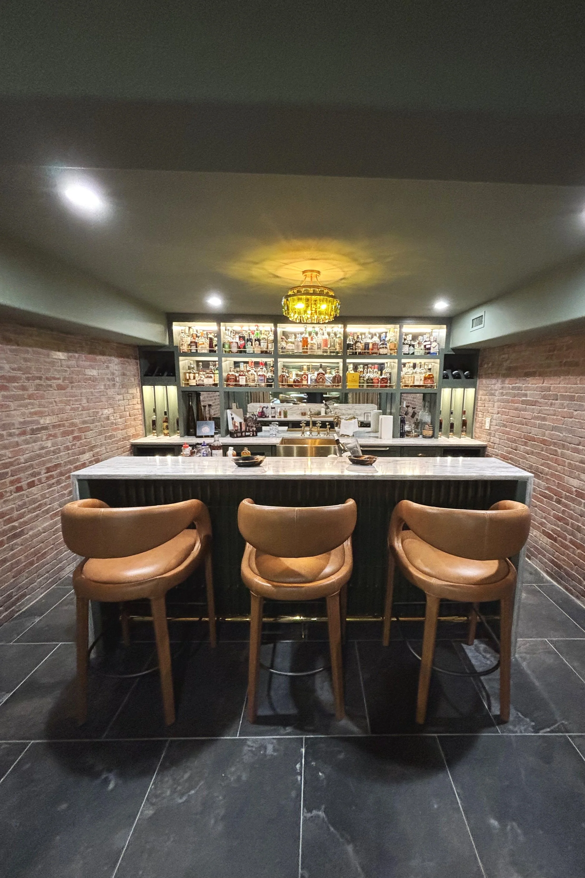 A bar with three brown leather chairs in front of a white marble countertop, with shelves of liquor bottles behind the bar in a cozy, dimly lit space with brick walls and black tiled floor.