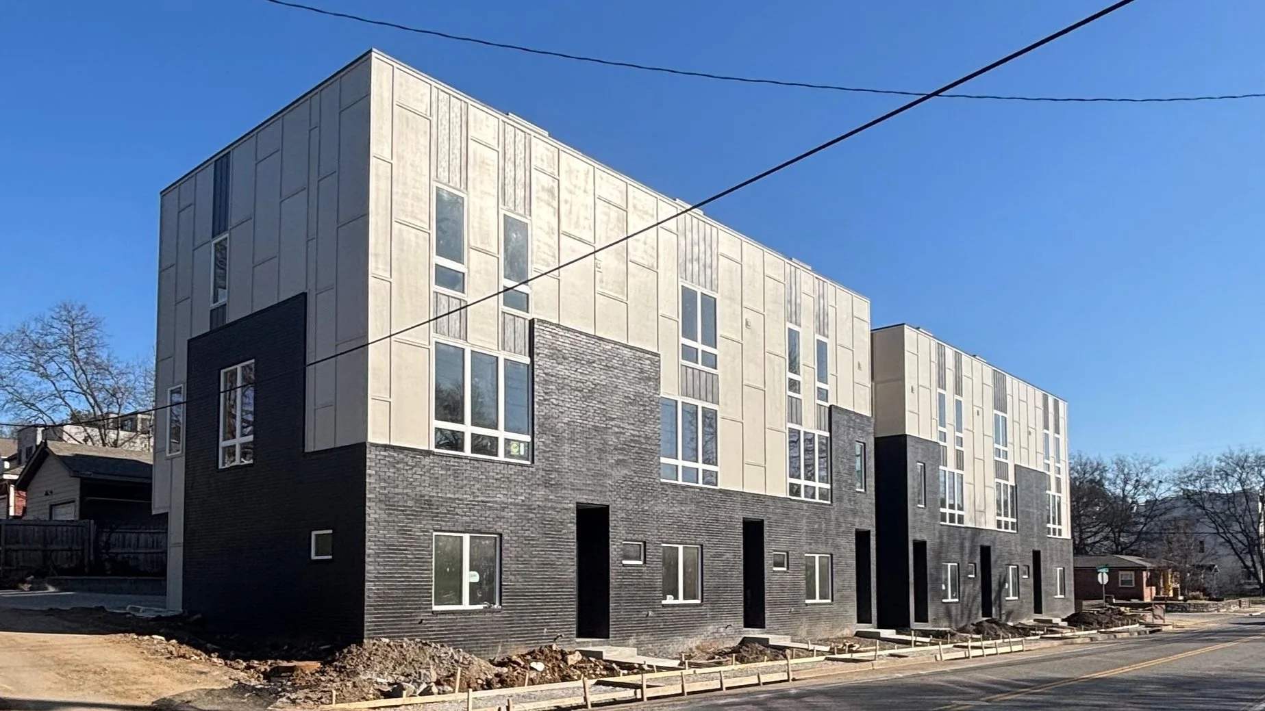 Modern multi-story residential building with a mix of black and beige exterior, large windows, and ongoing construction on the sidewalk in front.