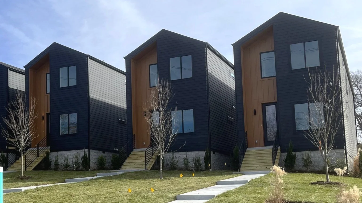 Three modern black and wood-paneled townhouses with small front yards, stairs leading to each front door, and leafless trees in the foreground under a cloudy sky.