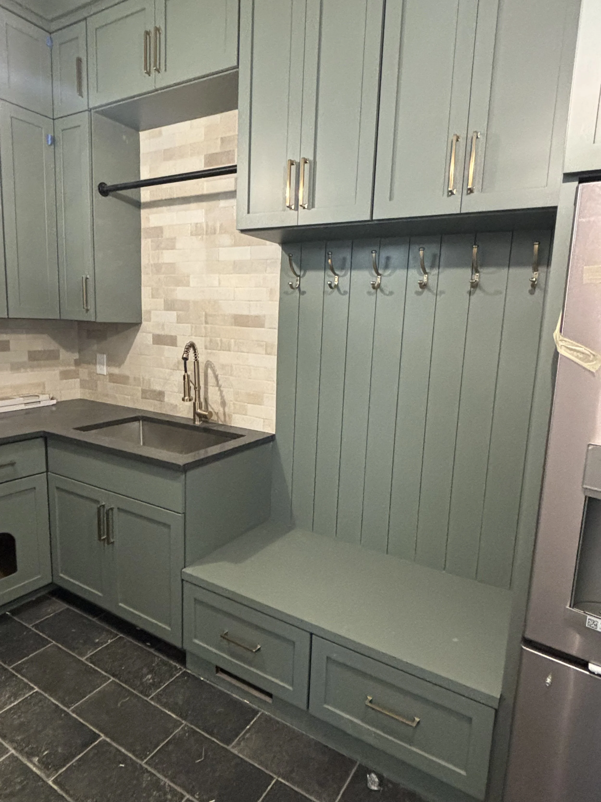 Kitchen with green cabinets, a black countertop, a sink with a gold faucet, and a tiled backsplash. There are hooks on the wall and drawers at the base of the cabinets.