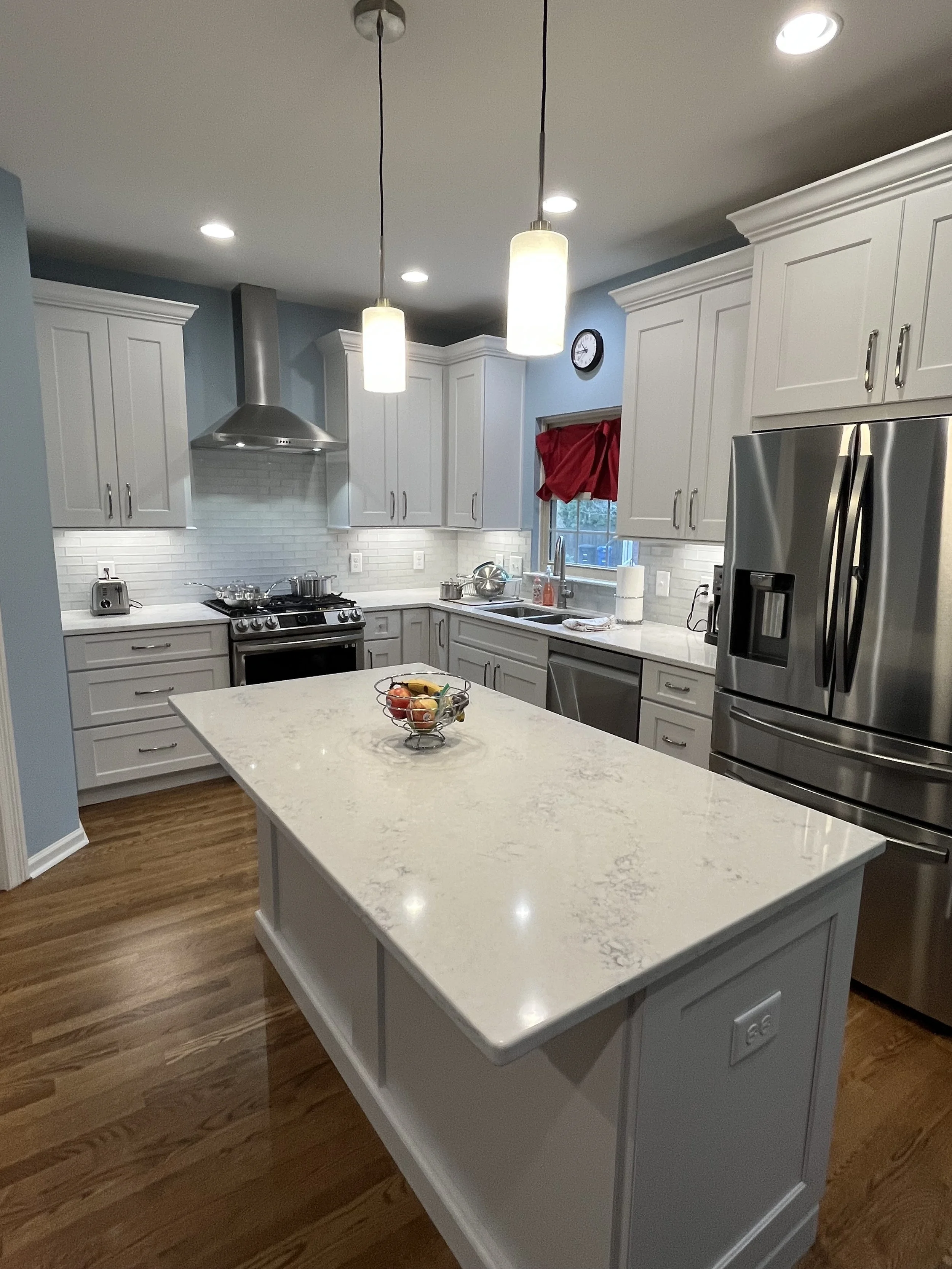 A modern kitchen with white cabinets, a marble kitchen island, stainless steel refrigerator, oven, and a window with a red curtain.