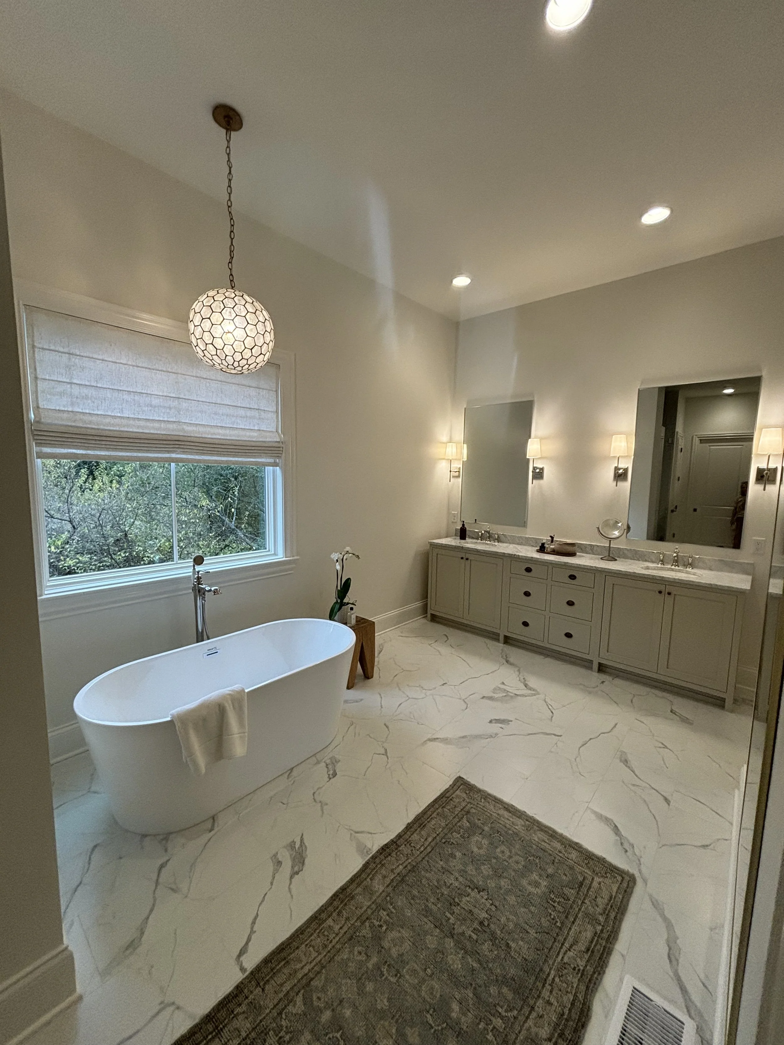 A spacious bathroom with a large window, a freestanding bathtub, a vanity with a mirror, and marble flooring.