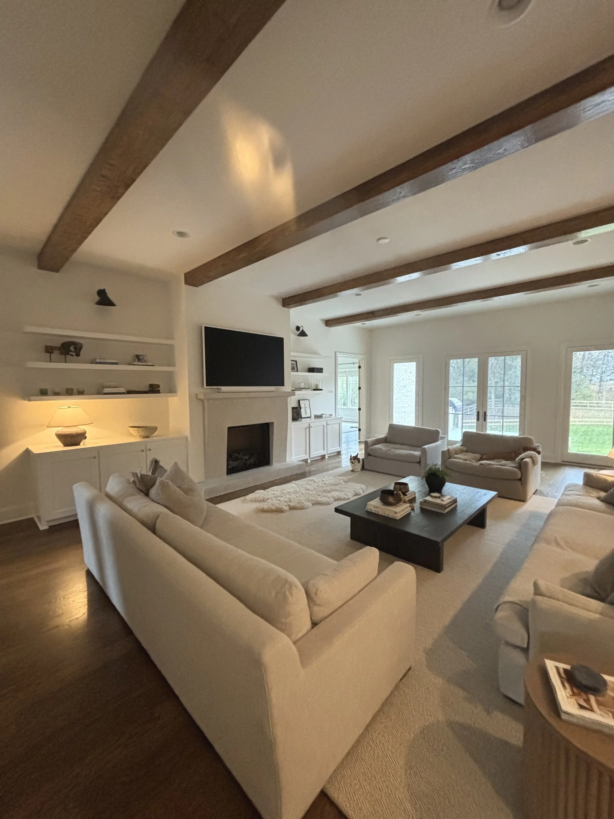 Living room with white sofas, a black coffee table, a fireplace, mounted TV, shelves, and large windows with a view of outdoor greenery.