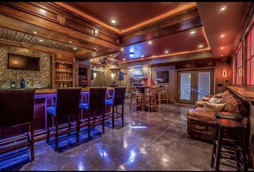 Home basement remodel in Nashville with a cozy bar with wooden fixtures, bar stools, a lounge area with leather sofa, and a small stage with a TV, illuminated by warm and colorful lighting.