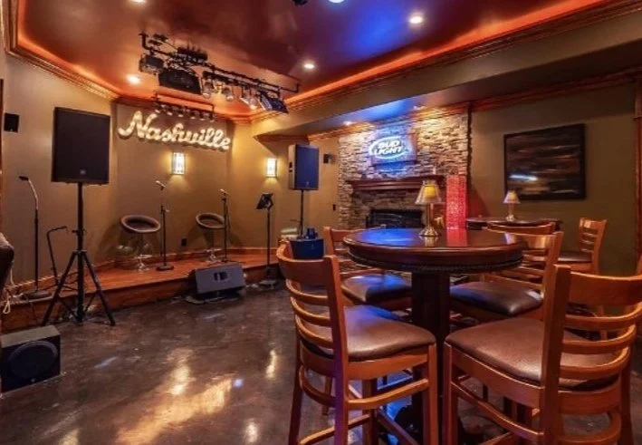 Interior of a small music venue or bar with a stage, musical instruments, and seating area. There is a sign that reads 'Nashville' and a neon sign that says 'Bud Light' on the brick wall.